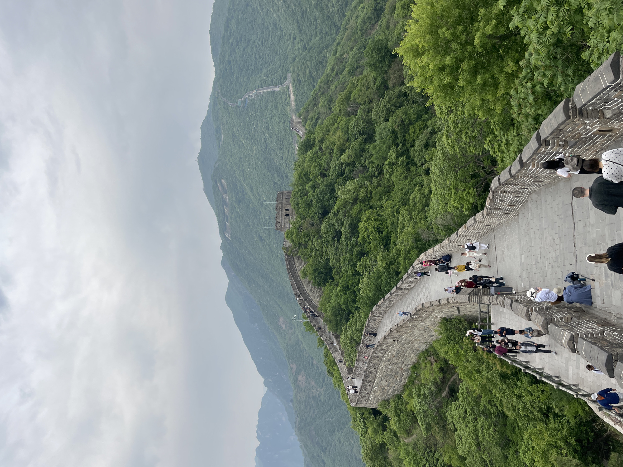 This photograph depicts the Great Wall of China, specifically a section located in the Huairou District of China. The image is taken from an elevated vantage point along the wall, looking along its course as it winds through a mountainous, tree-covered landscape. People are walking along the wall. The sky is overcast with a grey sky. The overall scene is a blend of natural beauty and man-made historical construction, portraying a popular tourist destination.
