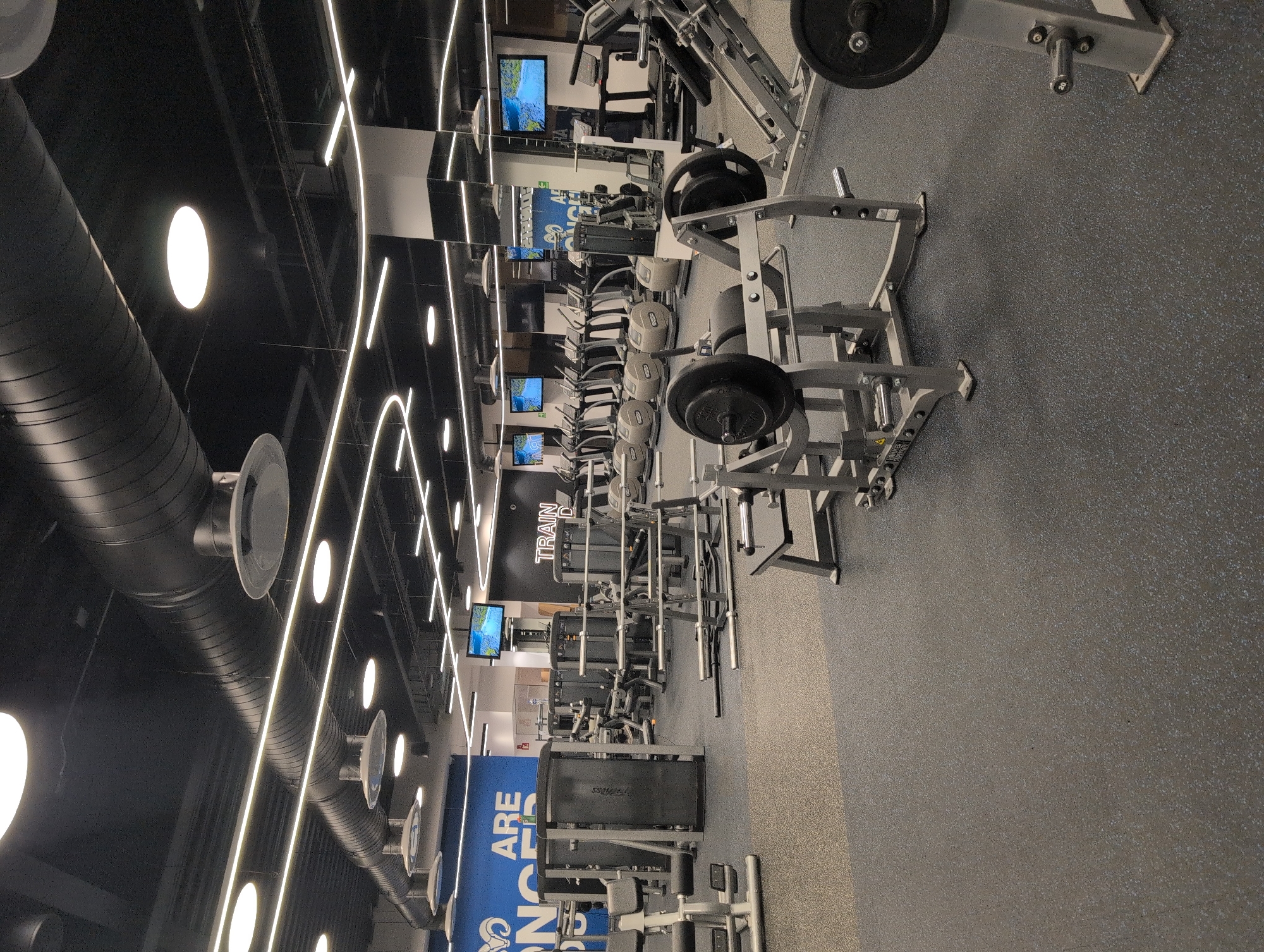 This is a full-shot, eye-level, medium-quality indoor photograph of a modern, well-lit gym. The gym features a variety of exercise equipment, including weight machines, treadmills, and ellipticals, arranged neatly on a grey rubberized floor. The ceiling is black with exposed ventilation ducts and numerous circular and linear lights. A large blue wall with white text reading "ARE STRONGER" is visible on the left, and another wall on the right has white text that says "TRAIN D". Multiple flat-screen televisions are mounted on the walls, displaying nature scenes. No people are present in the image. The environment appears clean and organized.