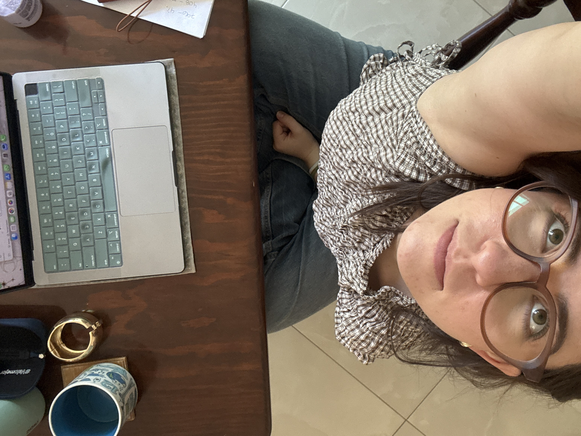 A person is seated at a dark wooden table, capturing a close-up, upward-facing self-portrait or being photographed. The individual is a female with dark hair, wearing brown-framed glasses and a white and brown plaid or gingham patterned top, with dark pants visible. She looks directly at the camera with a neutral to slightly contemplative expression.
On the table to her left, a silver Apple MacBook is open, with a light green/teal keyboard and a grey sleeve underneath. The screen is partially visible, showing app icons. Beside the laptop, a blue cap with the text "@Valomejor" is present, along with a gold-colored ring or bracelet and a blue and white patterned ceramic mug with a blue interior. In the upper right corner of the table, a piece of paper with handwritten notes, including calculations like "796-4%", "408", and "80h", is visible. The floor is covered in light-colored tiles, and part of a dark wooden chair arm is seen behind the person.
The setting appears to be an indoor, casual environment, possibly a home or personal workspace, in what is contextually identified as Puebla, Mexico. The lighting suggests daytime. The laptop features a QWERTY keyboard layout with visible keys like "command," "option," "shift," "caps lock," "delete," and "return," and the laptop sleeve has the brand "MOSISO" printed on it.