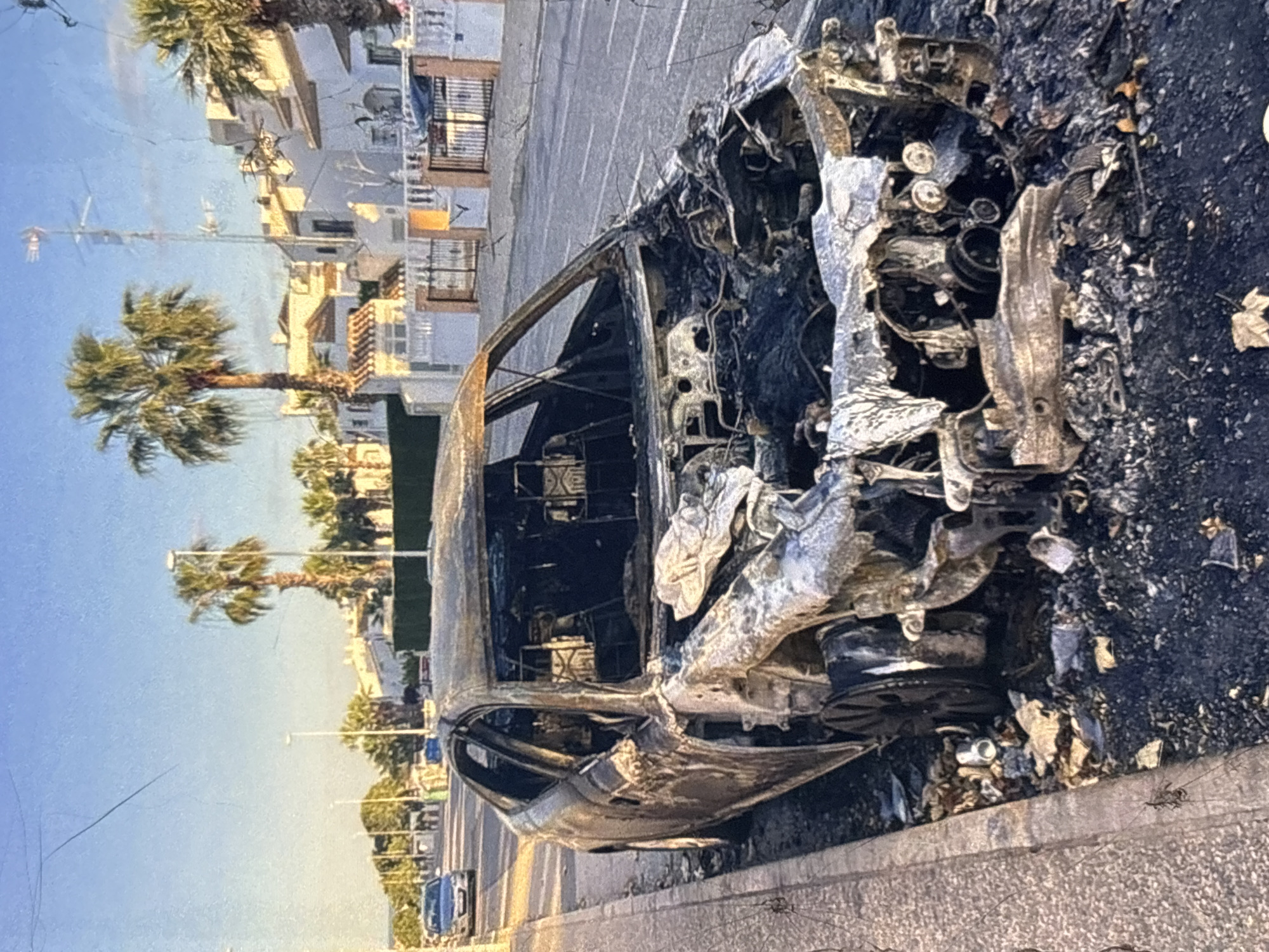 A severely burnt-out car is parked on a paved surface adjacent to a sidewalk. The car's interior is visible and extensively damaged by fire, with melted and charred components. The exterior also shows significant fire damage, with melted paint and distorted metal. The surrounding environment includes buildings with balconies and palm trees, suggesting an urban or suburban setting in a warm climate. The sky is clear and blue. Based on the visual cues, the location appears to be in the city of Orihuela, Spain, characterized by its architecture and vegetation.
