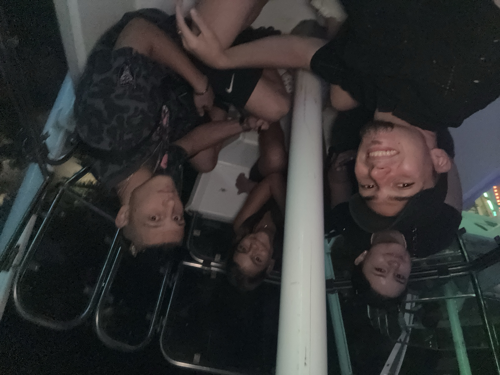 Four young adults are seated inside what appears to be a carnival ride at night. The ride is enclosed and dimly lit, with some colorful lights visible in the background. The people are positioned close together, suggesting they are friends or a couple enjoying an outing. The person in the foreground on the left is a young man wearing a black t-shirt and a black baseball cap, smiling broadly. To his right, and slightly behind him, is another young man wearing a dark, patterned t-shirt. Behind them and to the left, a young woman with dark hair is looking towards the camera. In the center background, another young woman with darker hair is visible, also looking towards the camera. The environment suggests a recreational setting, possibly a fair or amusement park. The image does not contain any information that would allow for identification of the specific location in Burgas, Bulgaria.