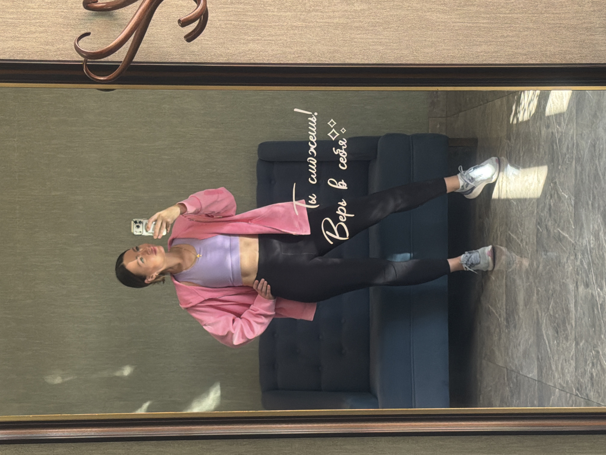A woman is taking a selfie in front of a mirror. She is wearing a lavender sports bra, black leggings, and a pink hoodie. The mirror has the text "Ты сможешь! Верь в себя" written on it. In the background, there is a gray couch and gray marble flooring. The location is possibly in Ovoshchnoy, Russia.
