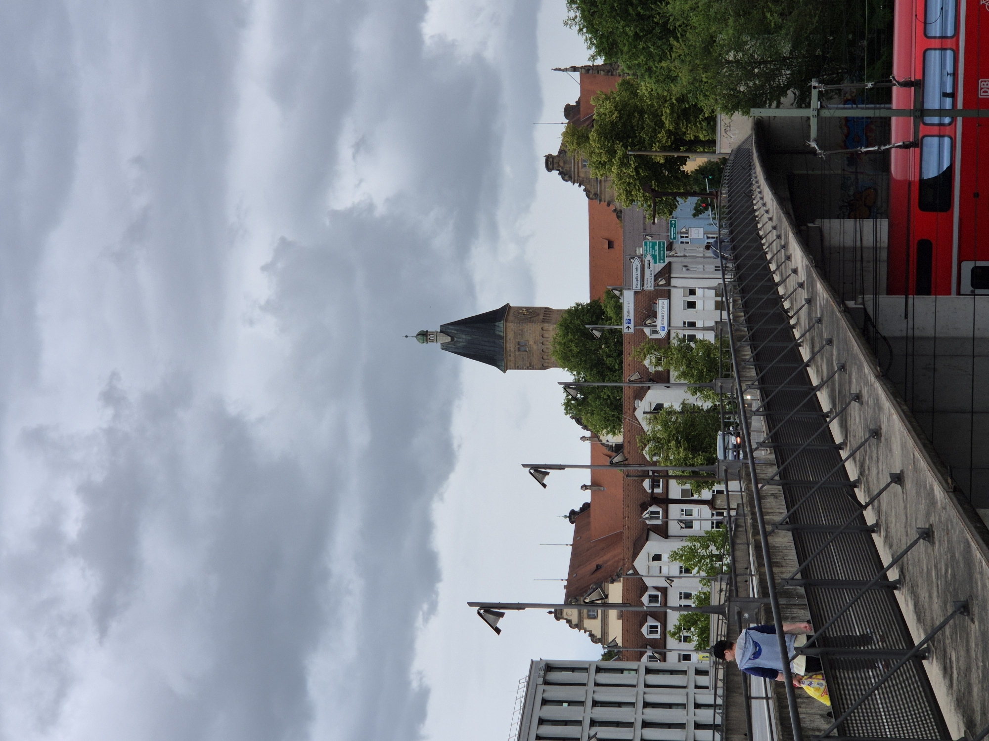 The image depicts a slightly overcast day in Regensburg, Germany. A pedestrian bridge crosses foreground to midground, with a person walking across it from left to right, carrying a yellow bag and wearing a blue jacket and black cap. The bridge overlooks a red train, possibly a DB Regio, passing below. In the background, various buildings with red-tiled roofs are visible, including a prominent tower with a dark, pointed roof and a clock face. Several street signs are also present. The environment suggests an urban setting with a mix of historical and modern architecture, and green trees dot the landscape.