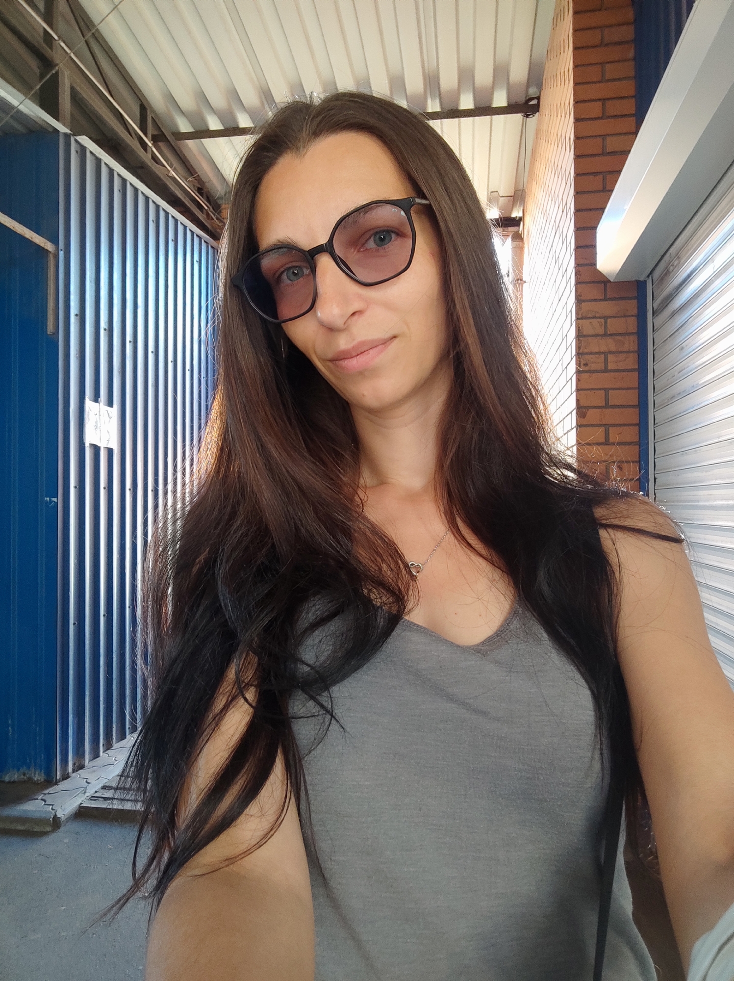 A woman with long, dark brown hair, wearing round-framed glasses and a grey tank top, takes a selfie. She has a small nose ring. The background shows a corrugated metal wall on the left, a brick wall on the right, and a corrugated metal roof overhead. The scene appears to be outdoors, possibly in a commercial or industrial area. The lighting suggests it is daytime. The context implies the location is the city of Kryvyi Rih, Ukraine.