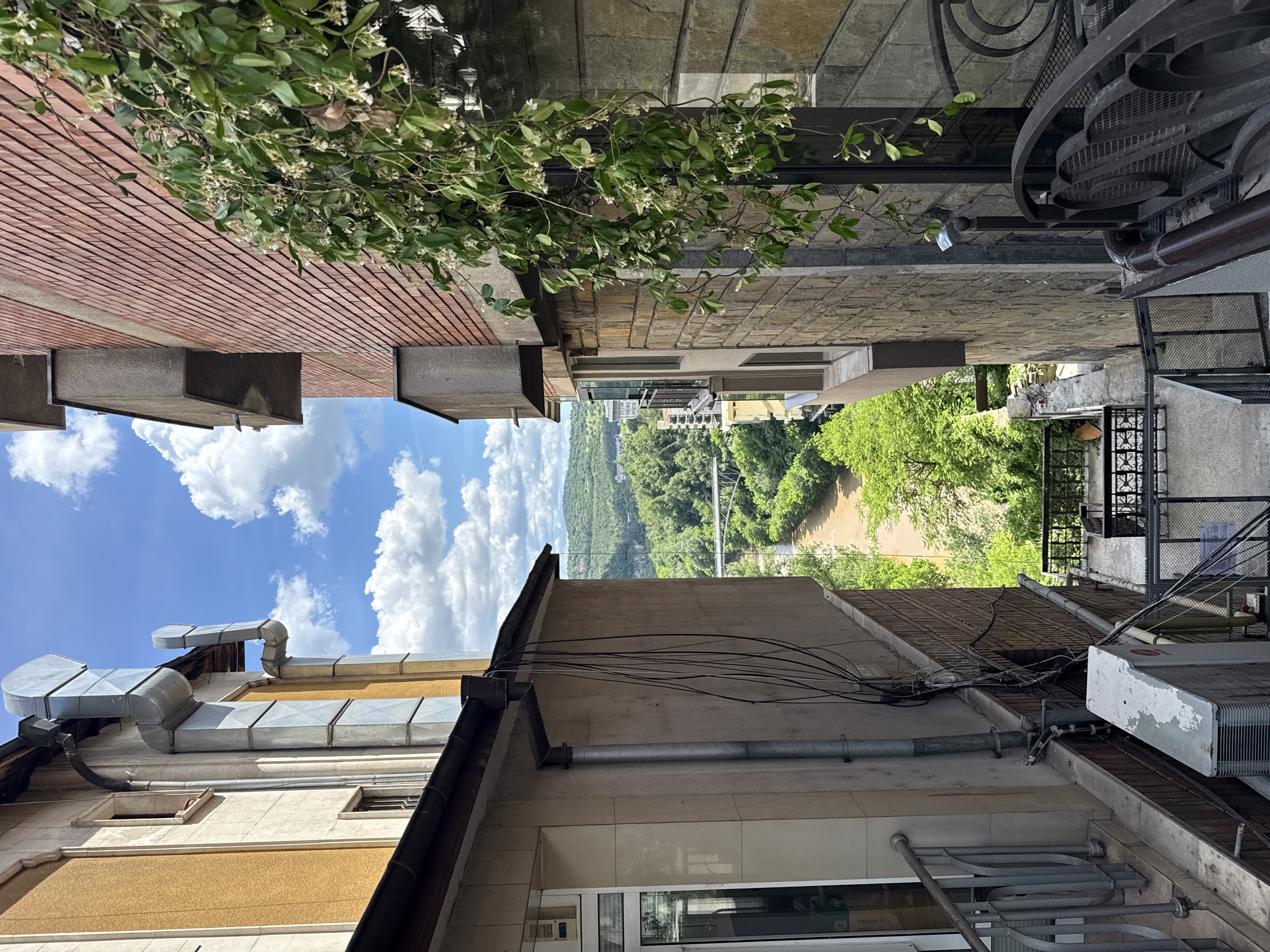 The image shows a view between buildings in Veliko Tarnovo, Bulgaria. Looking outward, a bridge crosses a river cutting through a forested area. The sky is blue with scattered white clouds. The foreground is dominated by the sides of adjacent buildings, showing architectural details like windows, air conditioning units, pipes, and a decorative wrought iron balcony. A brick building is visible on the left side of the image, with a climbing plant growing above.