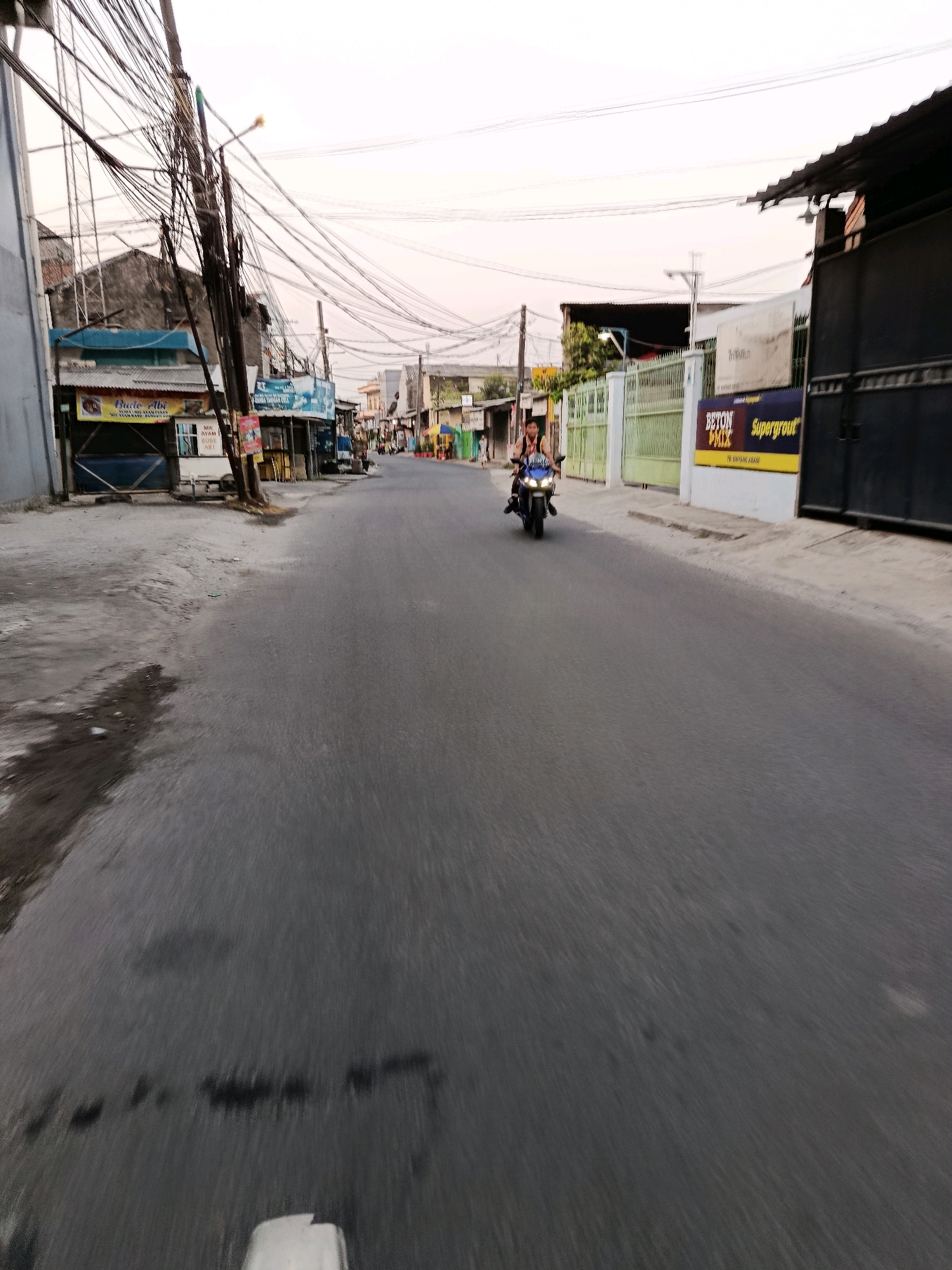 This is a street scene in Bekasi, Indonesia, likely taken during dusk. A person is riding a blue motorcycle down the asphalt road, which shows signs of wear and some damp patches. On the left side of the road, there are buildings with signage, including a blue structure with a yellow and red sign that appears to be for "Bude Abi" and "Mie Ayam." Further down the street on the left, there are more shops and overhead electrical wires are densely strung across utility poles, creating a complex network. On the right side of the road, a white building features a prominent yellow and black sign advertising "BETON MIX Supergrout." Beyond that, there is a dark metal gate and a green fence. The sky is overcast with a pale hue, suggesting the sun has set or is about to set. The overall impression is of a commercial or residential street in an urban area.