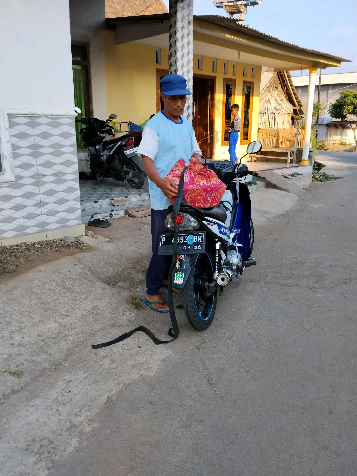 A man wearing a blue cap and a light blue t-shirt is standing next to a blue and white motorcycle parked on a concrete path. He is holding a red and gold patterned bag in his hands, which he is placing onto the motorcycle seat. A black strap is lying on the ground in front of the motorcycle. In the background, there is a house with a yellow wall and a corrugated iron roof. A young boy in a patterned shirt and blue pants is standing on the porch of the house. The scene is likely in Tlogosari, Indonesia, given the presence of motorcycles and typical housing styles.