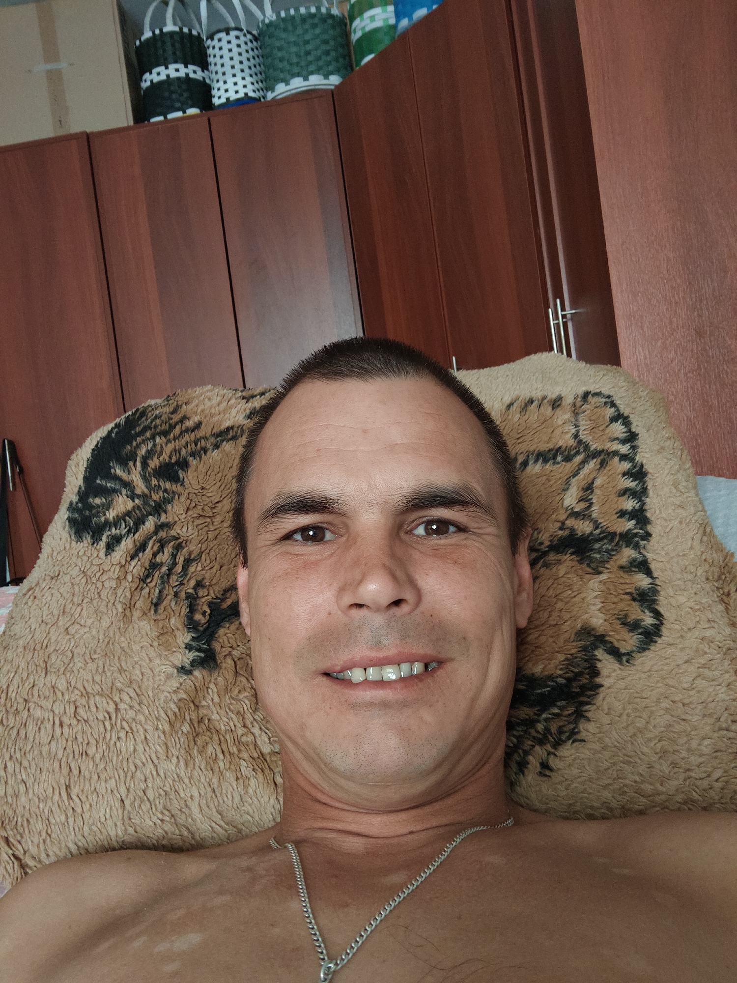 The image shows a close-up, selfie-style shot of a man lying down. He is shirtless and wearing a silver chain. He has short brown hair and is looking directly at the camera with a slight smile. Behind him, a patterned blanket with what appears to be a tiger print is visible. The background includes wooden cabinets and some wicker baskets on top of them. The overall impression is that the photo was taken indoors in a residential setting. The image does not provide enough information to determine a specific location within the city of Kurashim, Russia.