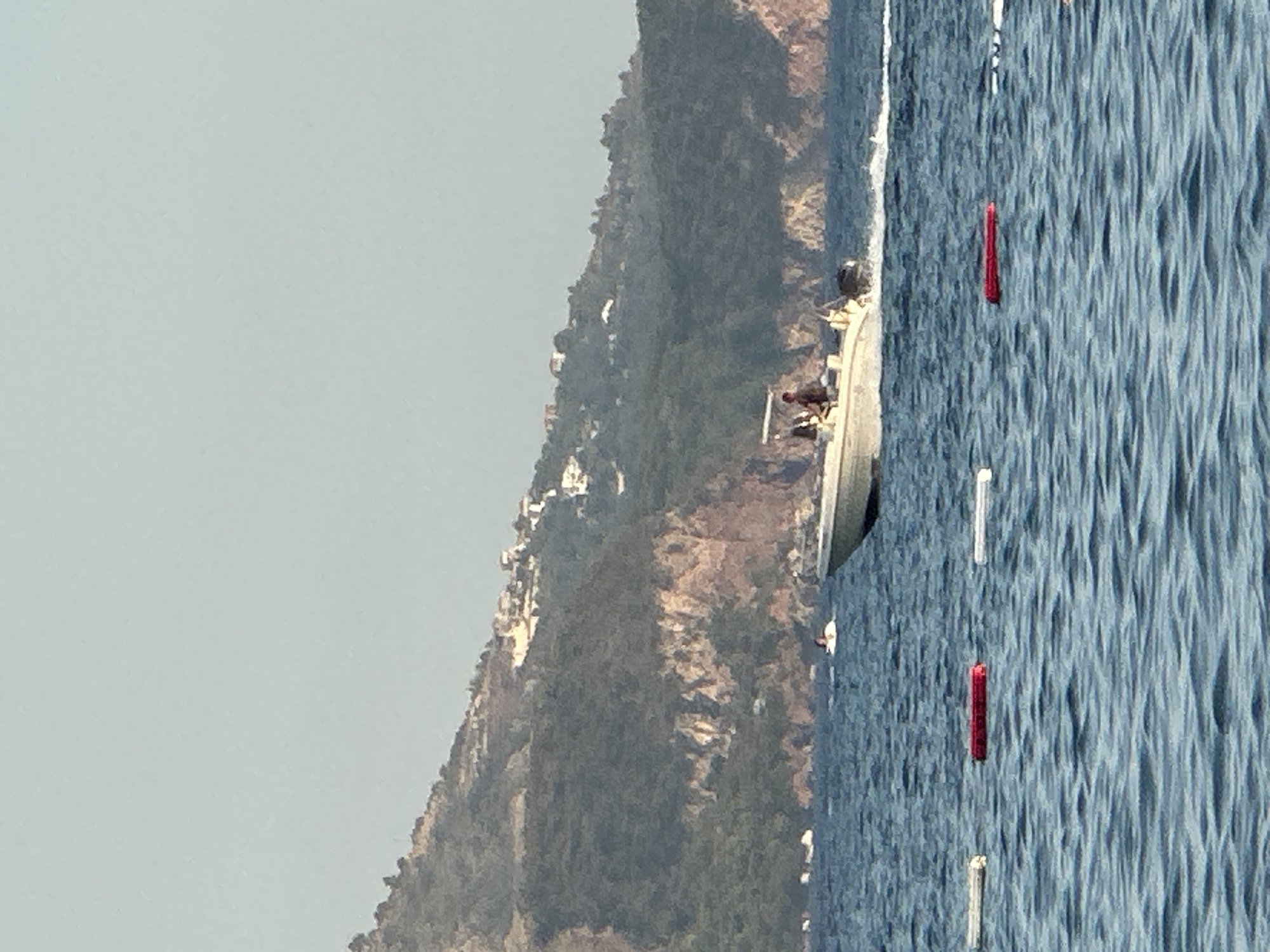 A white motorboat with two individuals aboard travels at speed through blue ocean waters. The boat is angled to the right, creating a spray of white water behind it. In the background, a densely vegetated hillside rises steeply from the water, dotted with white buildings. The sky above is a pale, hazy blue. The foreground shows the rippling surface of the water with several buoys, one red and two white, floating near the surface. The overall scene suggests a coastal setting near Boreti, Montenegro.