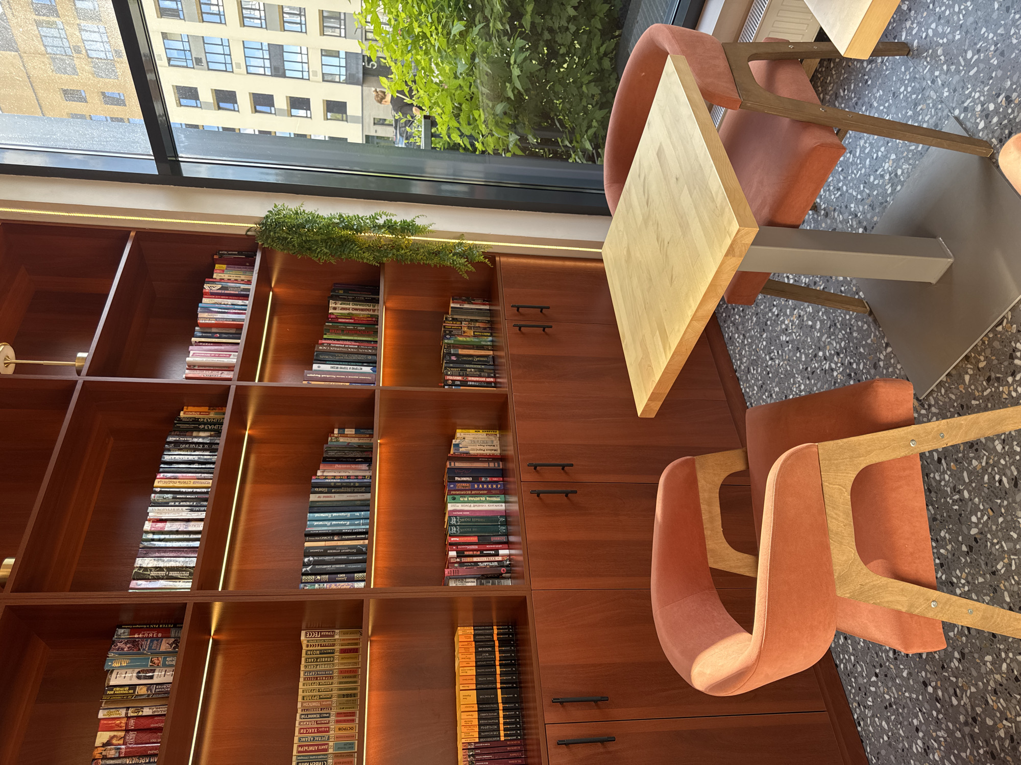 This is a photograph of a modern, well-lit indoor space that appears to be a library or study area within a building. The primary subjects are a large wooden bookshelf filled with books, two modern orange chairs, and a light wood table.
The bookshelf, made of dark wood, is organized into multiple rectangular shelves, each densely packed with books of various sizes and colors. Some shelves are illuminated by subtle LED strip lighting, creating an inviting atmosphere. A small green plant is placed on top of one section of the bookshelf.
The chairs are contemporary in design, with curved orange upholstered seats and backs, supported by light wood frames. One chair is placed at the table, while another is positioned to the side, its back facing the camera. The table has a light wood, angular top and a grey metal pedestal base.
In the background, a large window offers a view of an urban environment. A multi-story building with numerous windows is visible, along with lush green foliage, suggesting it is daytime and likely a pleasant day outdoors. The floor is a speckled terrazzo material.
The scene conveys a sense of quiet study or relaxation. The lighting is bright and even, indicating the photograph was taken during daylight hours. The visible text on the book spines includes titles and author names in Cyrillic script, suggesting the location is likely Russia. Specific visible text includes "ВАНКИР", "СТАЛИНГРАД", "ДАНТЕ АЛИГЬЕРИ", "ДУГЛАС АДАМС", "1984", and "ГЕРМАН ГЕСС".