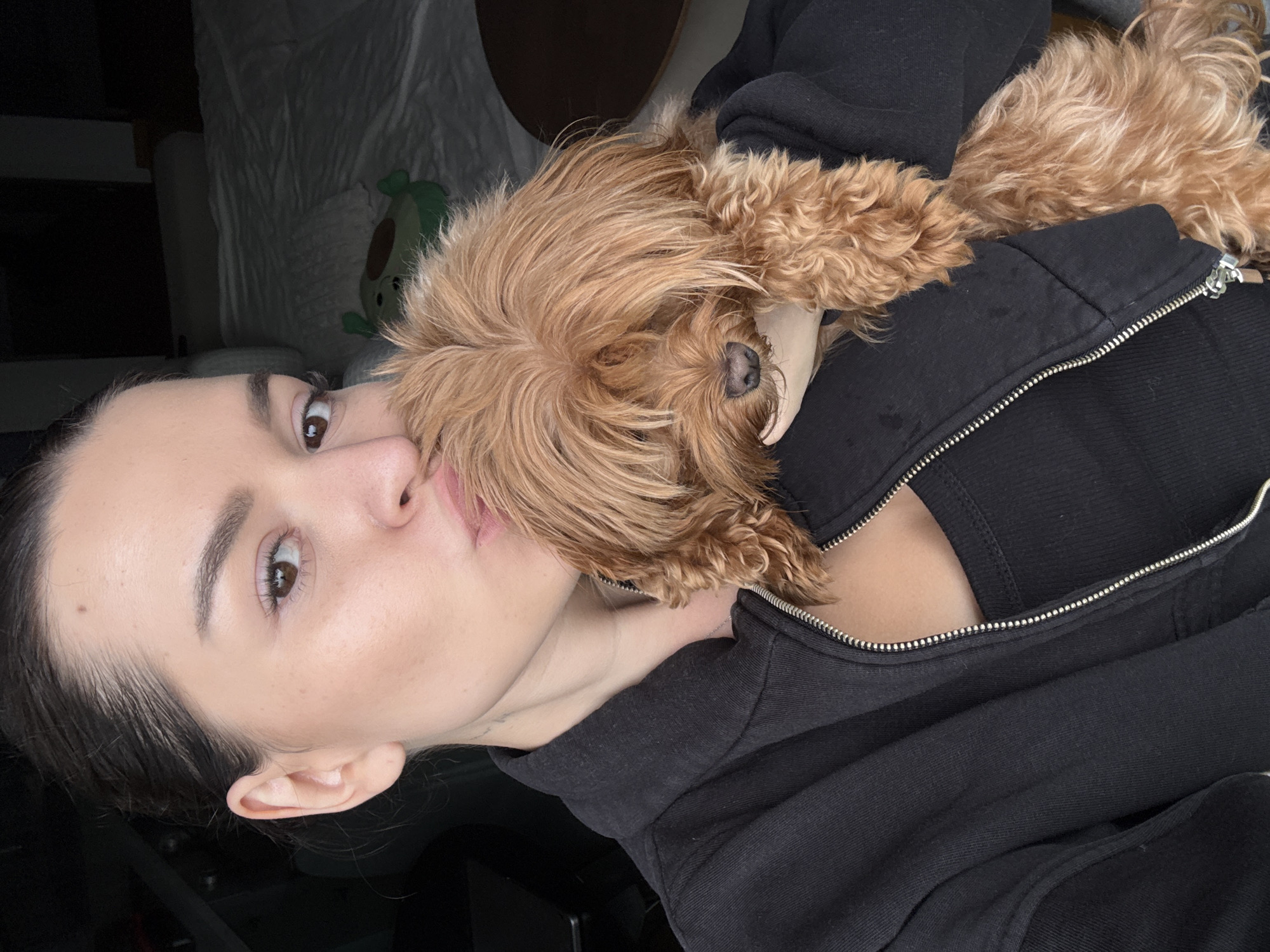 In a close-up, selfie-style shot, a young woman with dark hair pulled back into a bun is holding a small, tan-colored dog with shaggy fur. She is wearing a black zip-up hoodie over a black tank top and is leaning in to kiss the dog, whose face is mostly obscured by its fur. The background is dark and appears to be an indoor setting, possibly a bedroom, with a white-sheeted bed visible. There is a pillow with a green cartoon character on it on the bed. The image is underexposed in some areas, suggesting low lighting conditions. No specific location in Prague, Czechia is discernible from the image.