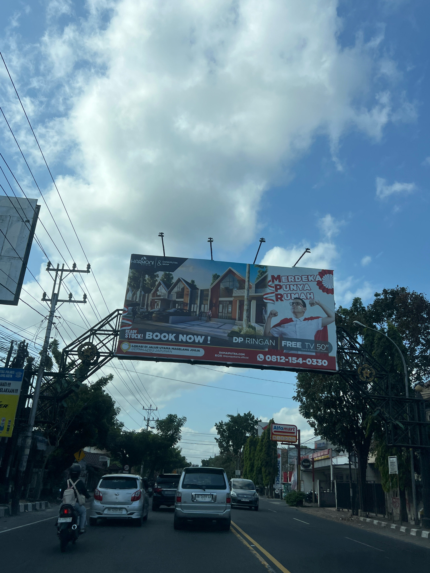 The image depicts a street scene in Magelang, Indonesia, on a clear day with a partially cloudy sky. A large billboard is prominently featured, advertising "HARMONI ESTATE & DAMAI PUTRA GROUP" properties with the slogan "MERDEKA PUNYA RUMAH" (Independence Has a Home). The billboard shows illustrations of modern houses and a person flexing their muscles in a red and white cape, suggesting a patriotic theme and a promotional offer of "DP RINGAN FREE TV 50"" (Light Down Payment, Free 50" TV). The location is further identified on the billboard as "LOKASI DI JALUR UTAMA MAGELANG JOGJA DEPAN SEBU JAPUNAN" (Location on the Main Magelang-Yogyakarta Route, in front of Sebuh Japun). The phone number 0812-154-0330 is also displayed.
In the foreground, traffic is visible on the road. A motorcycle with a rider wearing a helmet and backpack is in the left lane, followed by a silver car. Further ahead, a silver SUV and another vehicle are also on the road. The street is lined with trees and utility poles with overhead wires. On the right side of the road, a store with an "Alfamart" sign is visible, indicating a commercial area. The overall scene suggests a typical urban environment with ongoing traffic and commercial activity.