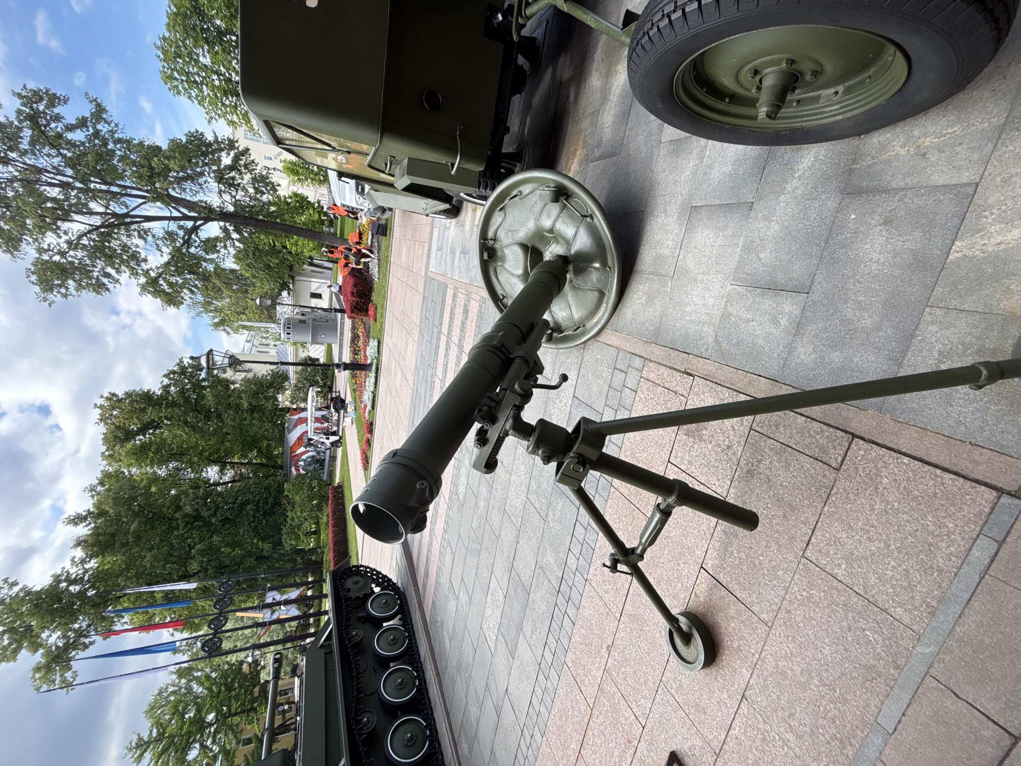 This image captures an outdoor display of military equipment, likely part of an exhibition or public event in Nizhny Novgorod, Russia. The primary subject in the foreground is a dark green mortar, resting on a sturdy tripod base with a circular ground plate, positioned on a light grey paved surface. To the right, part of a military vehicle is visible, showing a dark green wheel with a black tire and a section of its body. In the lower left foreground, the track system of another dark military vehicle, possibly a tank, is partially seen.
The scene unfolds in a public square or park, characterized by extensive light grey and light brown paving stones. Lush green trees with full foliage dominate the background, under a partly cloudy sky with patches of blue. Several traditional lampposts are scattered throughout the background. Further back, white or light-colored buildings with classical architectural elements are visible, suggesting an urban setting.
In the middle ground, alongside the paved path, there are vibrant flowerbeds with red and orange blooms. Several people, some wearing bright orange vests, can be observed in the distance, indicating event staff or visitors. Multiple flagpoles stand upright, displaying flags with colors including white, blue, and red, potentially Russian national or regional flags. The time of day appears to be daytime, with bright ambient light suggesting fair weather. There is no discernible text visible in the image.