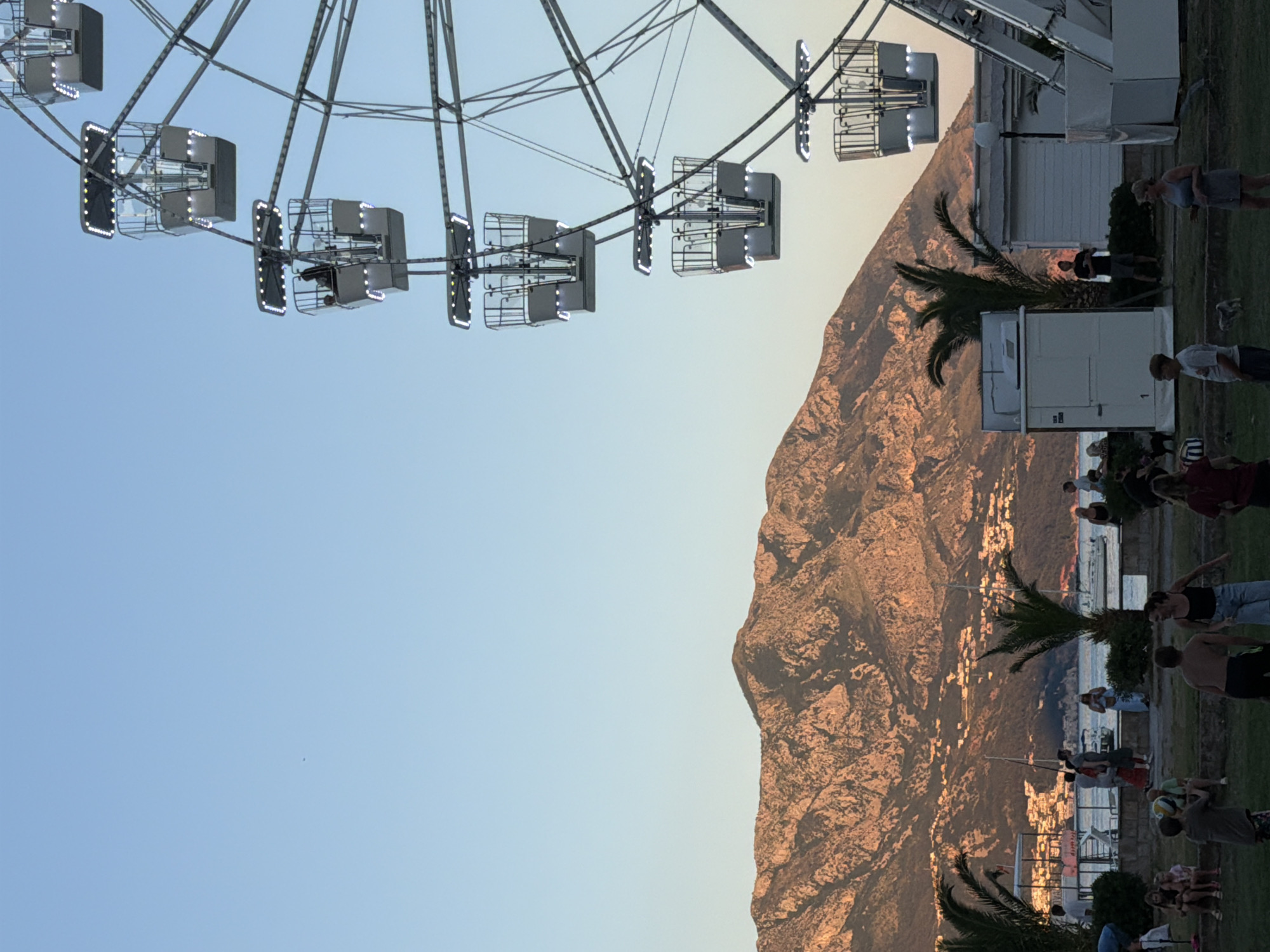 The image captures a scene at an amusement park near the city of Budva, Montenegro, during the golden hour. A Ferris wheel dominates the right side of the frame, with several illuminated gondolas visible against a clear blue sky. In the background, a mountain range bathed in the warm light of sunset forms a dramatic backdrop. The lower portion of the image reveals a grassy area with several people, including children and adults, engaged in various activities. Some are standing and talking, while others are playing with a ball. A few palm trees are scattered throughout the park, adding to the coastal atmosphere. In the mid-ground, there are several small white buildings and a body of water with boats, suggesting a seaside location. The overall mood is relaxed and festive, characteristic of a summer evening at a recreational area.
