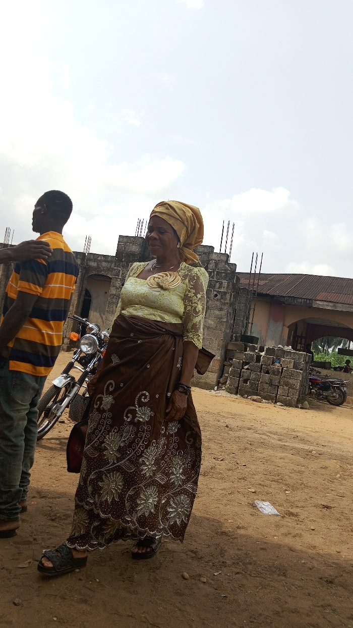This outdoor image captures a daytime scene in Agburu Uke, Nigeria, featuring two people on a dusty, unpaved ground under an overcast sky.
The central figure is an older woman, standing slightly angled with her gaze directed to her left. She wears a traditional yellow head wrap, a light green, short-sleeved lace blouse adorned with a spiral-shaped floral appliqué on the chest, and a long, flowing brown skirt intricately embroidered with silver or white floral and swirling patterns. She completes her attire with dark, open-toed sandals. Her expression appears calm or contemplative.
To her left, a man stands with his back mostly to the camera, his head turned slightly away. He is dressed in an orange and black striped short-sleeved shirt, blue jeans, and dark shoes. A hand, possibly belonging to another individual standing out of frame or his own, rests on his shoulder.
In the mid-ground, a silver and black motorcycle is parked near the man. The background features a structure built from concrete blocks, possibly under construction or in a state of disrepair, with exposed rebar protruding from the top. A building with a corrugated metal roof is visible to the right, and piles of stacked concrete blocks are scattered across the ground. A second, smaller motorcycle is discernible in the distant background on the right. The ground is dry, light brown dirt or sand. There is no visible text in the image.