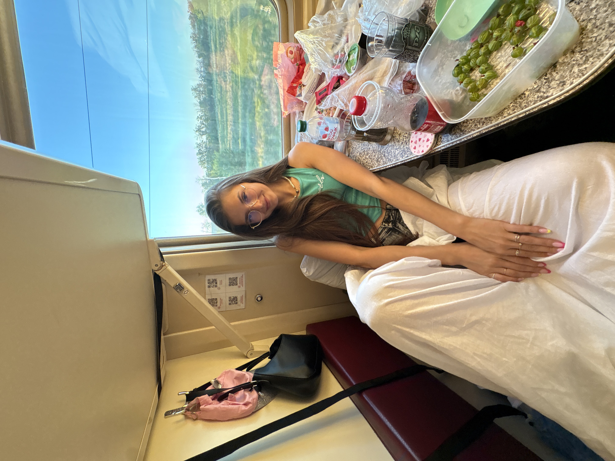 A young woman with long brown hair and glasses is seated in a train compartment. She is wearing a turquoise tank top and dark pants. A white blanket is draped over her legs. On the table in front of her are various food and drink items, including a container of green berries, bottles of water and soda, and bags of snacks. The train compartment appears to be in motion, with scenery visible through the window. The overall impression is of a casual journey. There is no indication of the city of Фисенково, Russia in the image.