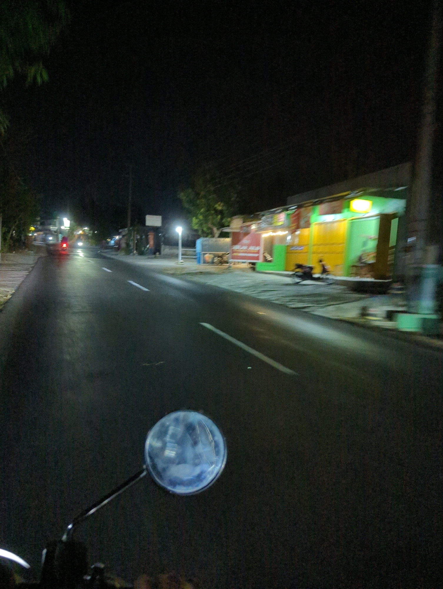The image depicts a night-time scene from the perspective of a motorcycle rider on a paved road in Kacangan, Indonesia. The road is wet and reflects the ambient light. On the right side of the road, a row of buildings with brightly colored facades, green and yellow, are visible. Some of these buildings appear to be shops or eateries, with illuminated signs and open doorways. A motorcycle is parked in front of one of the establishments, and two individuals are seated outside, possibly patrons. Streetlights illuminate parts of the scene, casting a glow on the road and the buildings. On the left side of the road, a sidewalk and some trees are present. The rider's rearview mirror is visible in the foreground, reflecting the dark surroundings. The overall impression is that of a moderately busy street at night in a local Indonesian town.