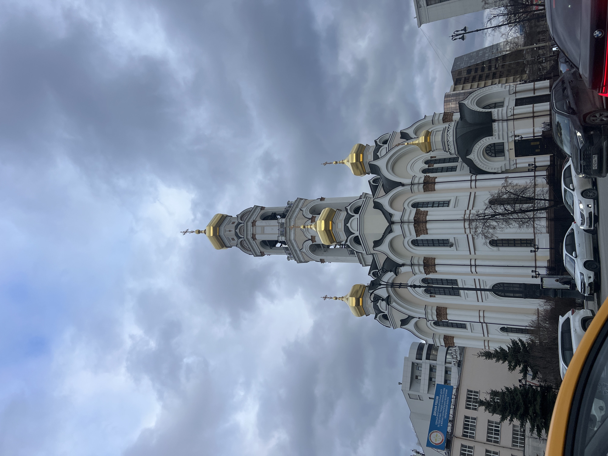 The image shows the Church on Blood in Yekaterinburg, Russia. It is a white and gold Russian Orthodox church with multiple domes and a bell tower, set against a cloudy sky. The lower part of the image shows a street scene with cars and a building with a sign "DOSAAF Russia". The image appears to be taken from within a car.