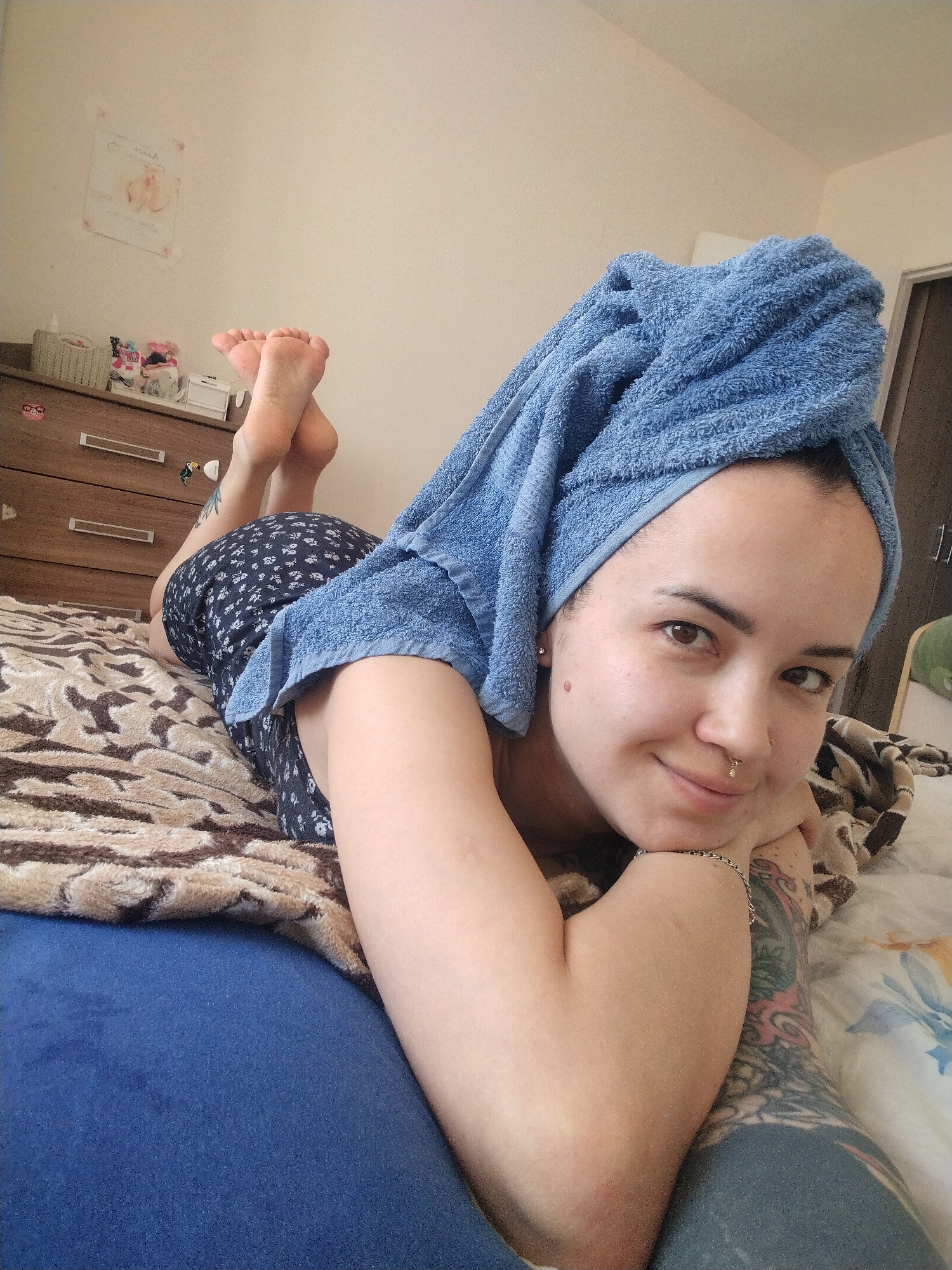 A woman is lying on a bed with her head propped up on her hands. She is wearing a blue towel wrapped around her head and a short, floral-patterned dress. Her feet are crossed at the ankles and raised slightly. She has tattoos on her right arm and a nose piercing. The bed has a brown and tan patterned blanket on top of a blue sheet. A wooden dresser is visible in the background, with various items on top. A small picture is hanging on the wall above the dresser. The location in the city of Noviy, Russia is not discernible.