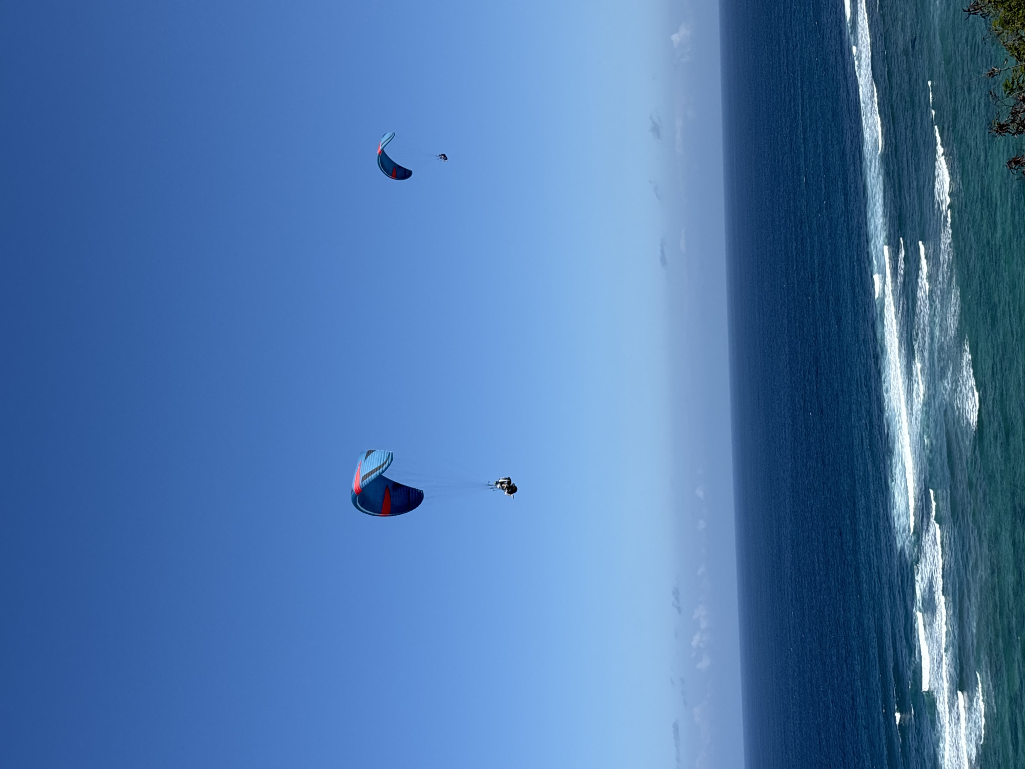 Two paragliders are in mid-air over the ocean. The paraglider in the foreground has a blue and red wing, with a person in a harness suspended below. The second paraglider, smaller and further in the background, also has a blue and red wing. The ocean below is a deep blue, with white waves breaking near the shore. The sky is clear and bright blue. The scene appears to be the coast of Kuta Selatan, Indonesia.