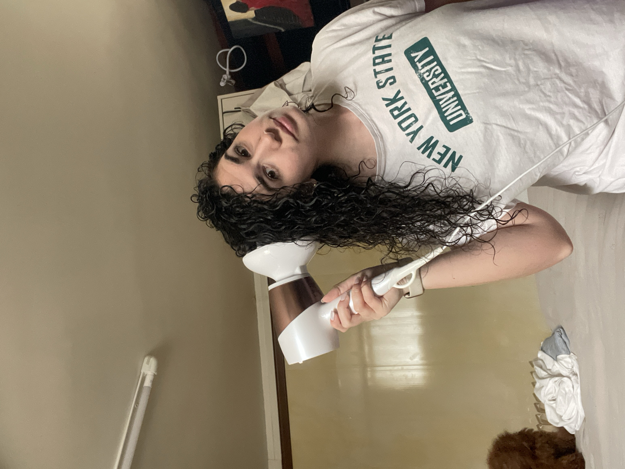 The image captures a young person, likely female, indoors in what appears to be Sofia, Bulgaria, in a candid moment while drying their hair. The person is positioned with their head tilted, seemingly bending over to dry their long, dark, and visibly wet curly hair with a hairdryer. They are looking directly at the camera with a neutral expression.
The subject is wearing a white short-sleeved t-shirt with green block letters prominently displaying "NEW YORK STATE UNIVERSITY". They hold a white and rose gold (or copper) hairdryer in their right hand, directing it towards their hair. A light-colored wristband or watch is visible on the same wrist.
The background primarily consists of a plain, light beige or light brown wall. On the left side, a segment of a white door frame or vertical trim is visible. In the upper right corner, some blurry, indistinct dark objects and a white cable or cord can be seen. Towards the bottom right, a bundle of white and grey cloth or laundry is resting on a surface. A white fluorescent light tube or similar fixture is partially visible in the bottom-left area of the frame. The setting appears to be an ordinary indoor space under artificial lighting, with no specific time of day indicated. The image itself is oriented sideways, suggesting it might have been taken from an unusual angle or rotated during capture.