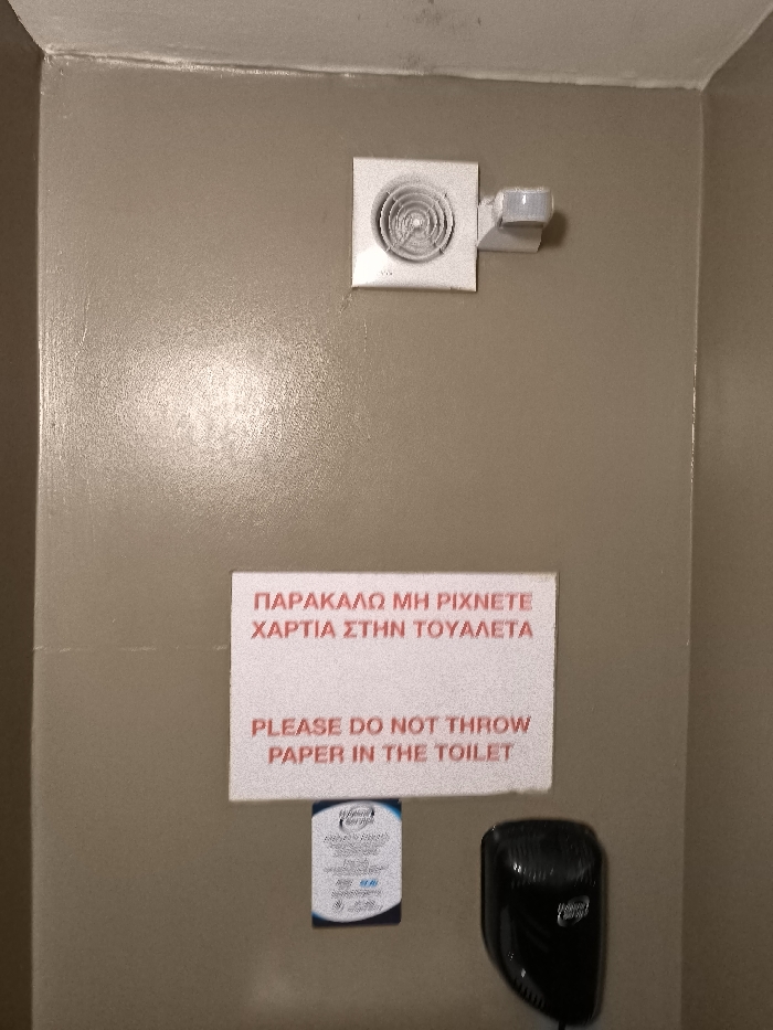 The image captures a beige-painted wall in what appears to be a public restroom or toilet facility, given the context of the signage. Mounted high on the wall is a white square exhaust fan with a circular grille, and to its right, a small white motion sensor.
Below these fixtures, centrally placed on the wall, is a white rectangular sign with red lettering. The sign displays an instruction in two languages. The top text is in Greek, reading: "ΠΑΡΑΚΑΛΩ ΜΗ ΡΙΧΝΕΤΕ ΧΑΡΤΙΑ ΣΤΗΝ ΤΟΥΑΛΕΤΑ". Beneath this, the English translation states: "PLEASE DO NOT THROW PAPER IN THE TOILET". This type of instruction is common in areas like Greece where plumbing systems may not be designed to handle toilet paper.
Further down and to the left of the hand dryer, a small, laminated card is affixed to the wall, possibly detailing hygiene services or similar information. The words "Hygiene Services" are partially visible on it. To the right of this card, a black, wall-mounted hand dryer is installed, its textured surface visible. The overall setting is utilitarian and functional. No people or specific activities are depicted; the image focuses on the installed fixtures and instructional signs within this space. The wall has a slightly glossy finish, reflecting some light.