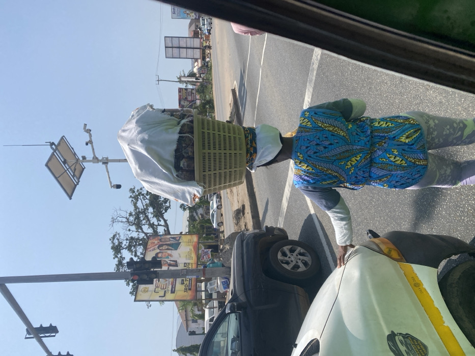 The image depicts a daytime street scene at an intersection in Adenta, Greater Accra Region, Ghana, under clear, sunny skies. In the immediate foreground, from a slightly elevated perspective within a vehicle, the rear of a woman is visible. She is a street vendor, balancing a large, light-green plastic woven basket filled with pineapples (partially covered by a white cloth) on her head. She wears a blue and yellow patterned sleeveless top over a light grey long-sleeved shirt and light grey patterned leggings, with a white headwrap. Her right hand rests on the hood of a light-colored vehicle, which shows a yellow stripe and minor wear, and has a black sticker on its fender. To the left of the vendor, a black pickup truck is stopped in traffic.
Further into the scene, a traffic light pole displays a red signal. Adjacent to it, a tall metal pole supports a solar panel array, an antenna, and multiple CCTV cameras, indicating a sophisticated urban monitoring system. A large billboard, predominantly yellow, advertises "NEXT LEVEL THE PEOPLE'S CHURCH" in Adenta, with specific dates "3RD - 7TH FEB." The road is paved asphalt with visible white lane markings. Several other vehicles are discernible in the distance, indicating active traffic. Trees and low-rise buildings are visible in the background, lining the roadside.