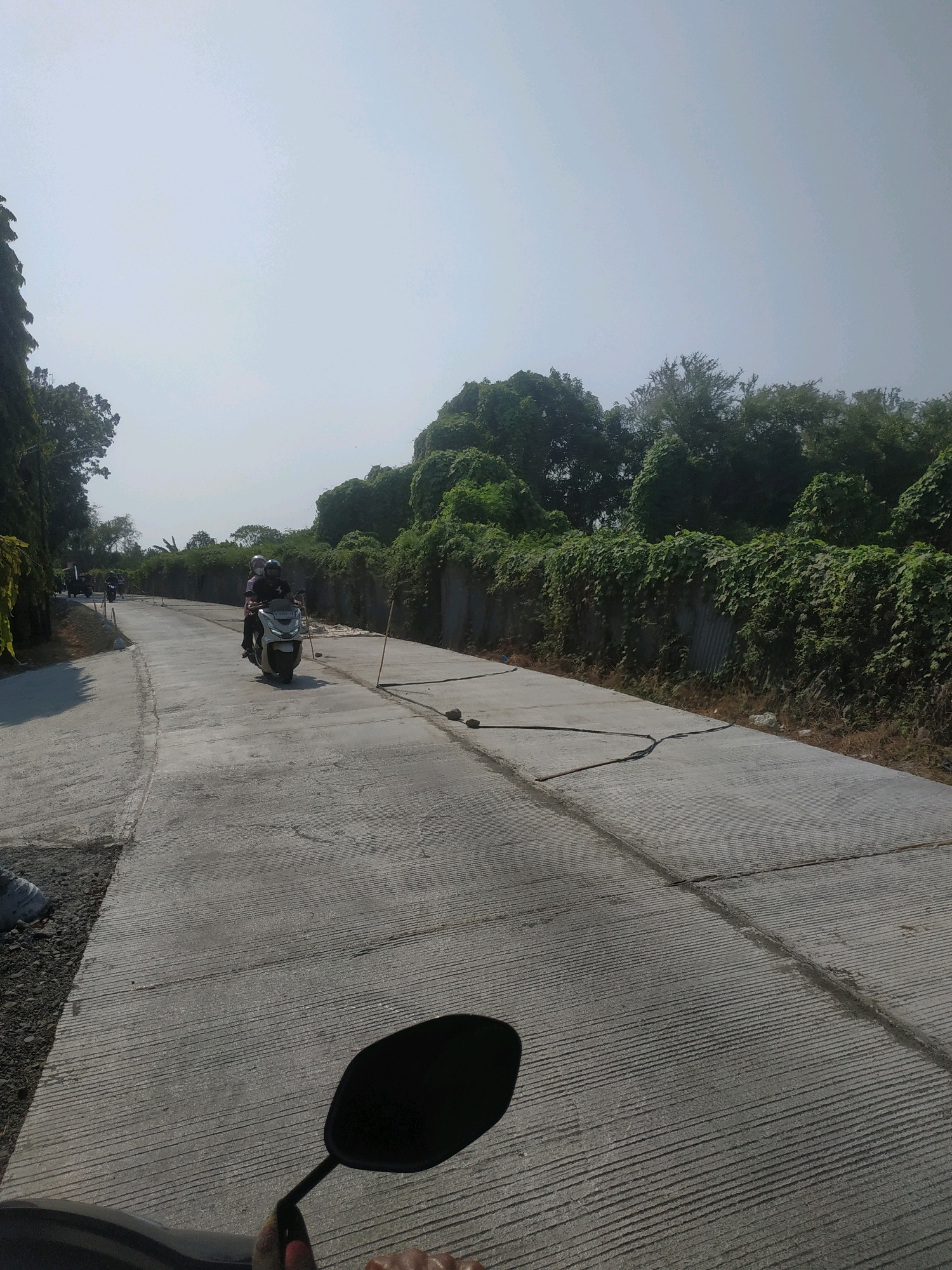 A drone perspective captures a partially paved concrete road curving through a semi-urban landscape in Mejasem Timur, Indonesia. On the road, two individuals on a white scooter are traveling away from the viewer. The road is flanked by a low, green-leafed vegetation wall on the right and a line of trees on the left. The sky is clear and bright, indicating daytime. The overall scene depicts a road under construction or recently completed, surrounded by lush greenery.