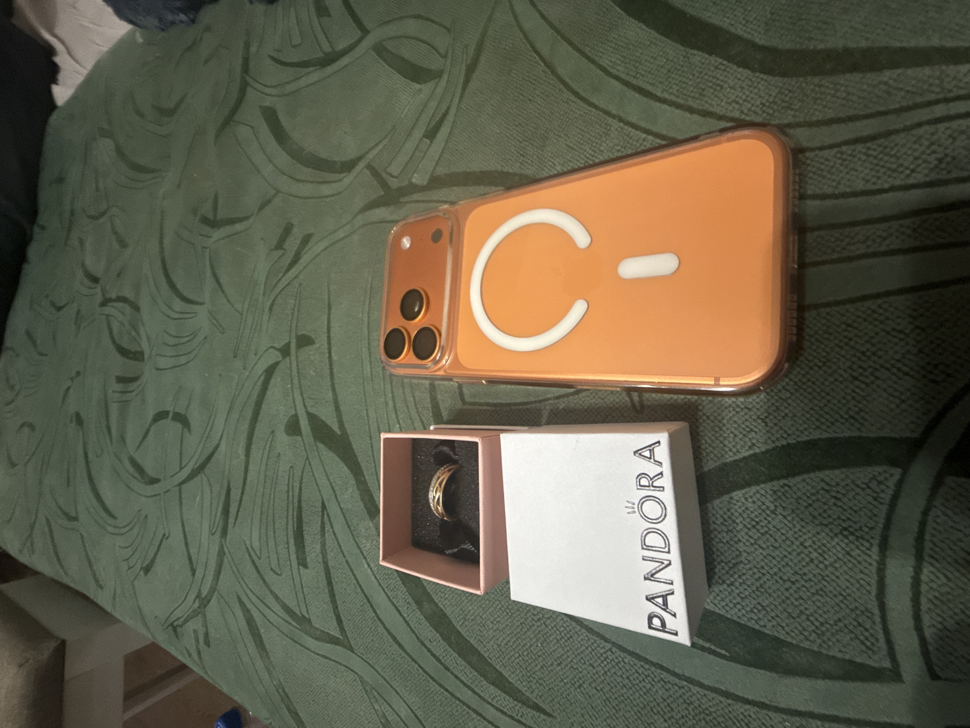 In an indoor scene, an orange phone case and a Pandora ring box are placed on a green, textured surface. The phone case is transparent with an orange tint and features a white magnetic ring and a small white rectangle on the back. The Pandora box is white with the brand's logo printed in black. Inside the box, a ring with a gold band and small stones is visible. The overall lighting suggests an indoor setting, possibly a bedroom. There are no people or animals present in the image.
