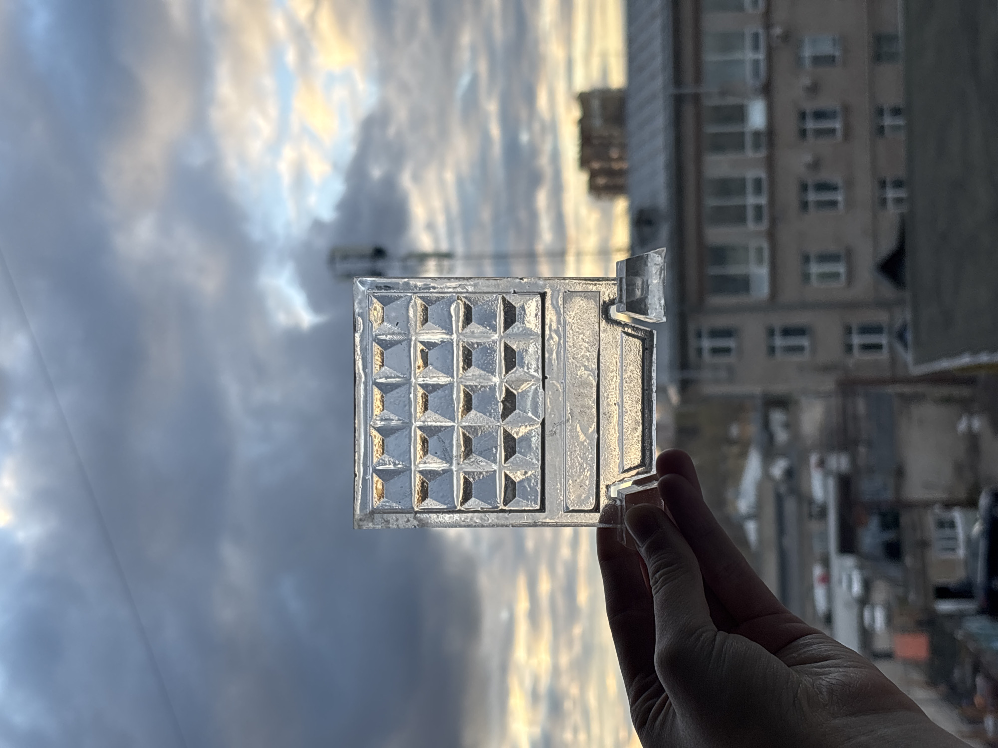 A hand holds a square glass object with a textured, grid-like pattern against a blurred background of a cityscape and sky. The grid pattern consists of smaller square sections with raised, pyramid-like centers. To the right of the patterned area, there's a smooth, elongated section and a small, detached piece extending outward. The hand holding the glass object is silhouetted, and only a portion of it is visible in the bottom right corner. The cityscape in the background appears to consist of apartment buildings with numerous windows, and the sky above is filled with clouds, illuminated by the setting sun. The location is Perm, Russia.