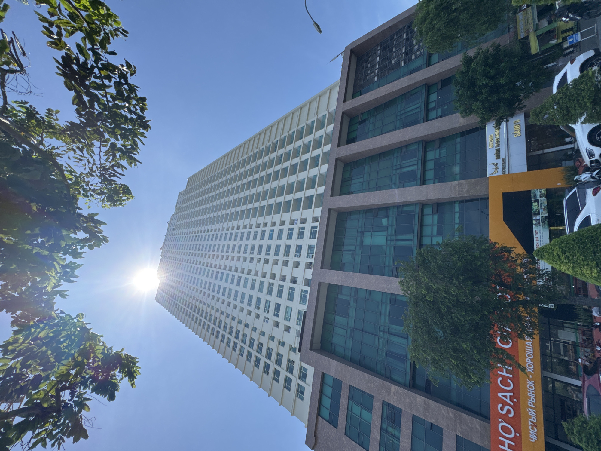 The image shows a low-angle, wide shot of a modern, tall, cream-colored apartment building against a clear blue sky with a bright sun peeking through green tree branches in the foreground. The building has a distinct podium structure at its base, featuring dark green tinted windows and a mottled brown facade. The apartment building itself has numerous rectangular windows and balconies. Below the apartment building, on the ground level, there are storefronts with orange and yellow signage. One sign displays text in Vietnamese and Russian, reading "HỌ SÁCH C" and "ЧИСТЫЙ РЫНОК - ХОРОШАЯ", which translates to "Bookstore" and "Clean Market - Good". Another sign visible is for "SANDI". Several vehicles, including cars and motorcycles, are parked on the street in front of the building. Trees and foliage are visible in the lower portion of the image. The overall scene suggests an urban environment in Nha Trang, Vietnam, likely a mixed-use development with residential and commercial spaces.