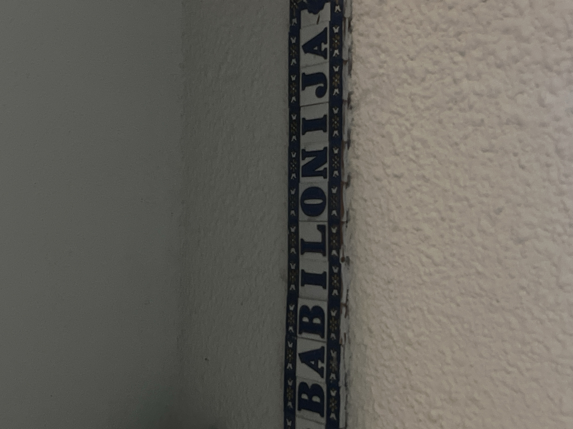 The image shows a vertical strip with the word "BABILONIJA" spelled out in blue capital letters, likely a sign or decorative element. The background consists of two walls, one smooth and dark grey on the left and the other textured and light beige on the right. The word "BABILONIJA" is the name of a shop or establishment in the city of Budva, Montenegro.