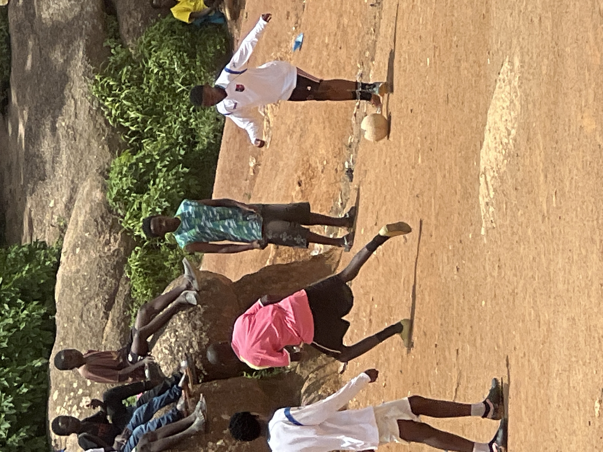 In Jos, Nigeria, a group of young boys are playing soccer on a dirt field next to a rocky outcrop covered in lush green vegetation. The atmosphere is casual and playful as they engage in a friendly match. Some boys are actively participating in the game, while others are observing from the sidelines, perched on the rocks. The scene captures the vibrant energy of youth in a rural Nigerian setting.
