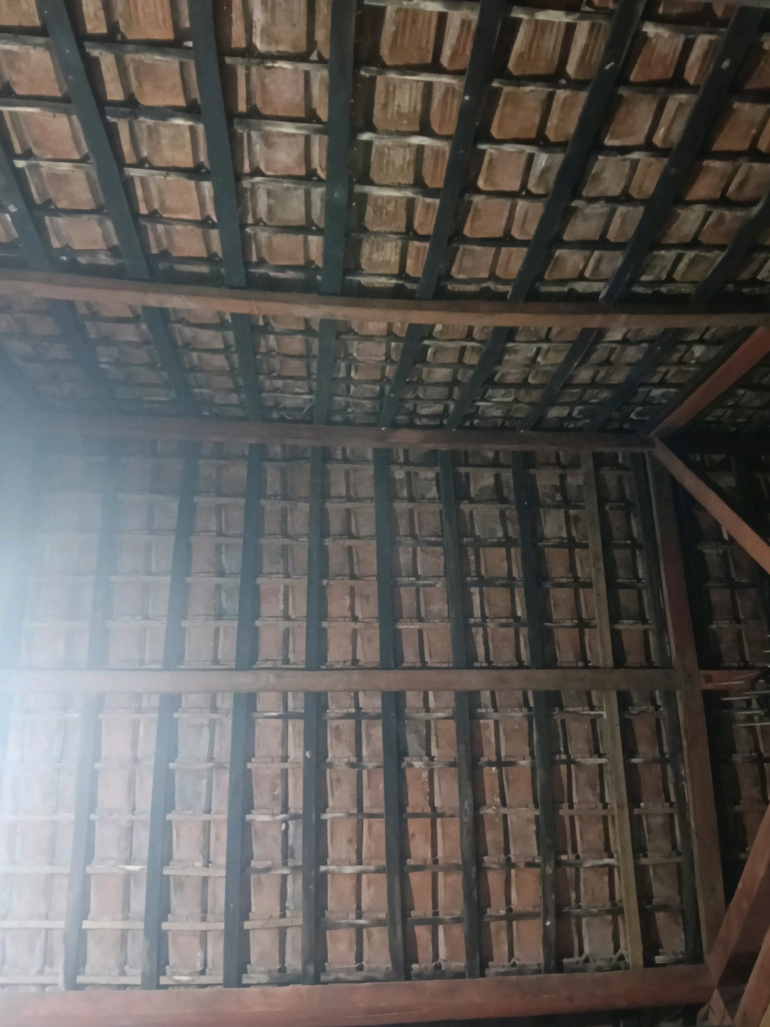 The image shows an underside view of a traditional tiled roof structure, possibly in a rural or older urban setting within Galuk, Indonesia. The roof is composed of overlapping terracotta tiles supported by a wooden framework. Dark, possibly wooden or metal beams run horizontally and vertically, creating a grid-like pattern beneath the tiles. Some of the beams appear to be stained or painted dark. In the upper portion of the image, the wooden beams are more prominent and form a more complex structure, suggesting the roof's peak or main supports. Light is entering from the left side, creating a bright glare that obscures some details of the tiles and beams in that area. The overall impression is of a rustic and possibly aged construction, characteristic of some architectural styles found in Indonesia. There are no people or animals visible in the image, and no objects or actions are depicted other than the static structure of the roof.