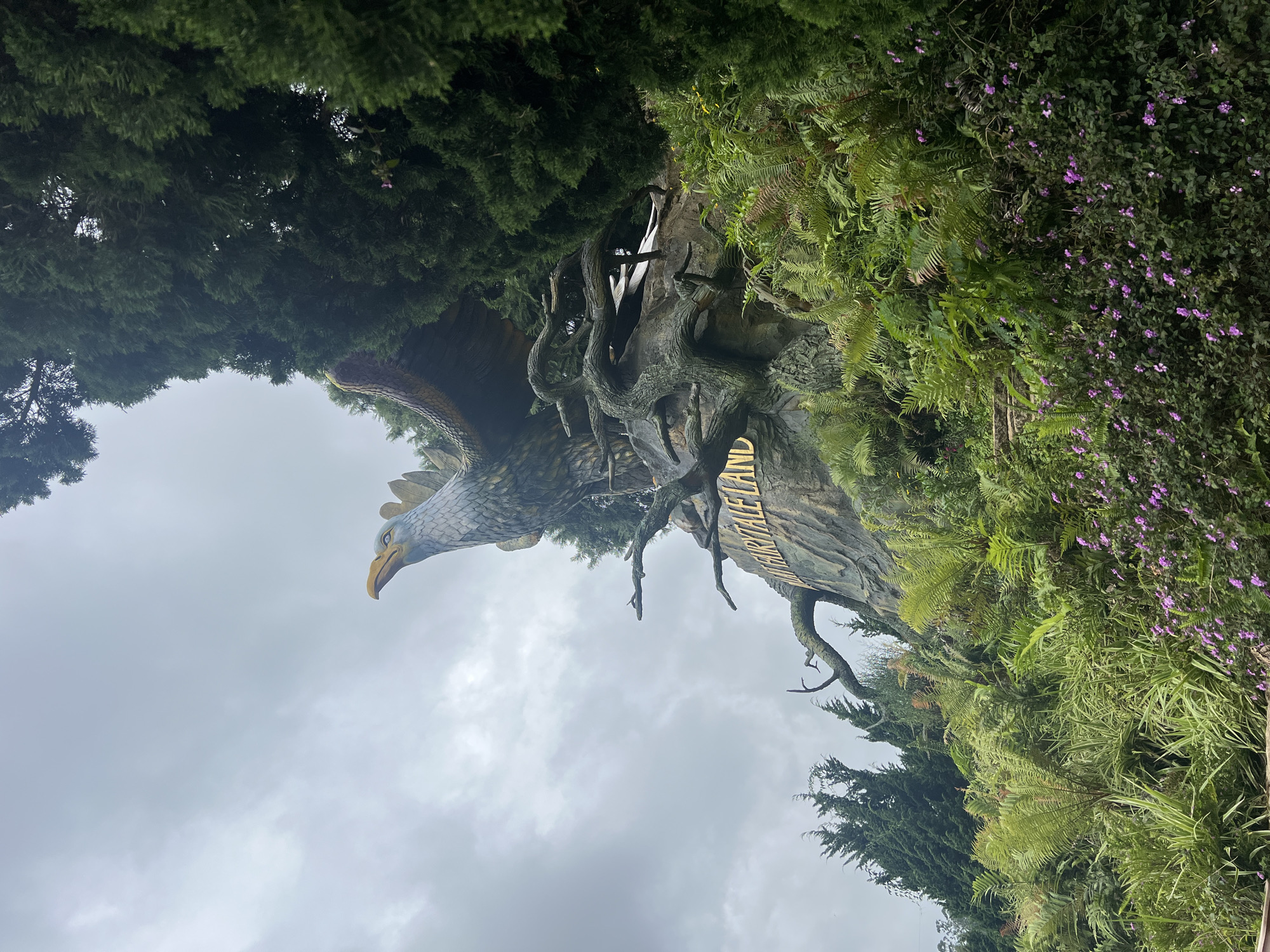 The image depicts a large, ornate statue of a mythological bird, possibly a phoenix or griffin, perched on a rocky outcropping. The statue is intricately detailed with feathers and a sharp beak. In front of the statue, a sign with golden lettering reads "FAIRYTALE LAND." The statue and the sign are surrounded by lush, green vegetation, including ferns and trees, with some small purple flowers in the foreground. The sky is overcast and cloudy. This scene is located at Da Lat Fairytale Land, an amusement park in Da Lat, Vietnam. There are no people visible in the image.