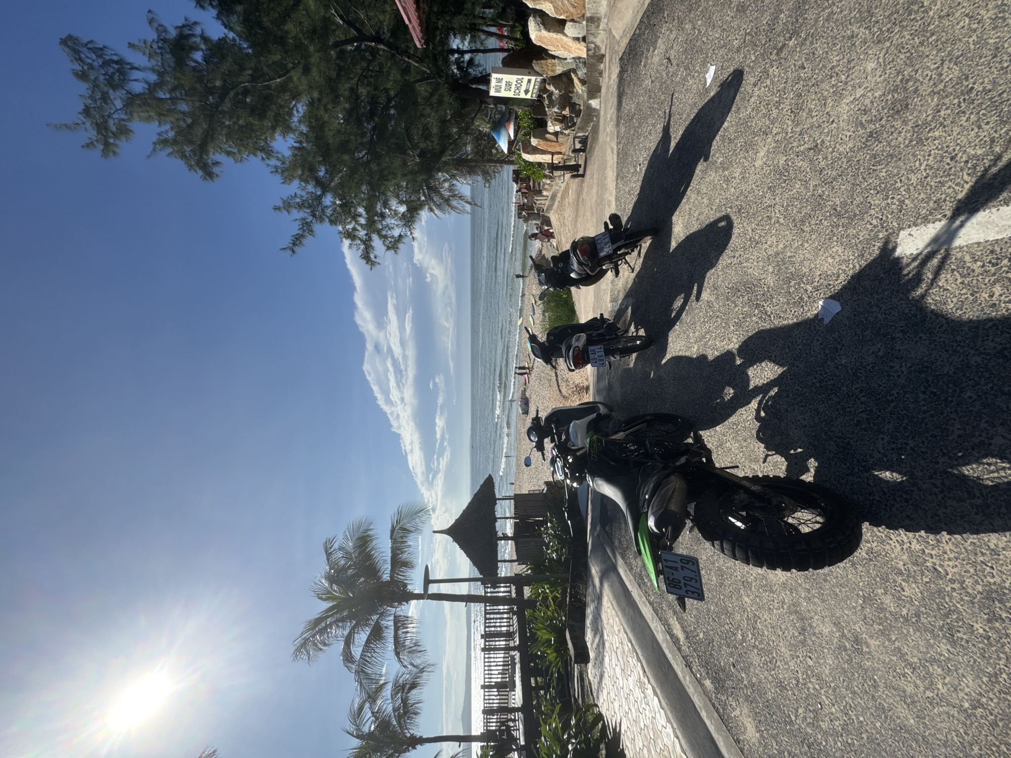 The image captures a sunny day at a beachside location, likely in Phan Thiết, Vietnam, given the presence of a "MŨI NÉ SURF SCHOOL" sign. In the foreground, three motorcycles are parked on an asphalt road that slopes gently towards the beach. The closest motorcycle is a green and black dirt bike with a license plate reading "86-A1 379.79." Behind it are two smaller black scooters. Palm trees line the left side of the road, and a traditional thatched-roof hut is visible further down. The beach is populated with a few people, and surfboards are scattered near the water. The ocean is calm with moderate waves, and the sun is shining brightly in a clear blue sky with some scattered clouds. The overall scene suggests a relaxed, tropical vacation atmosphere.