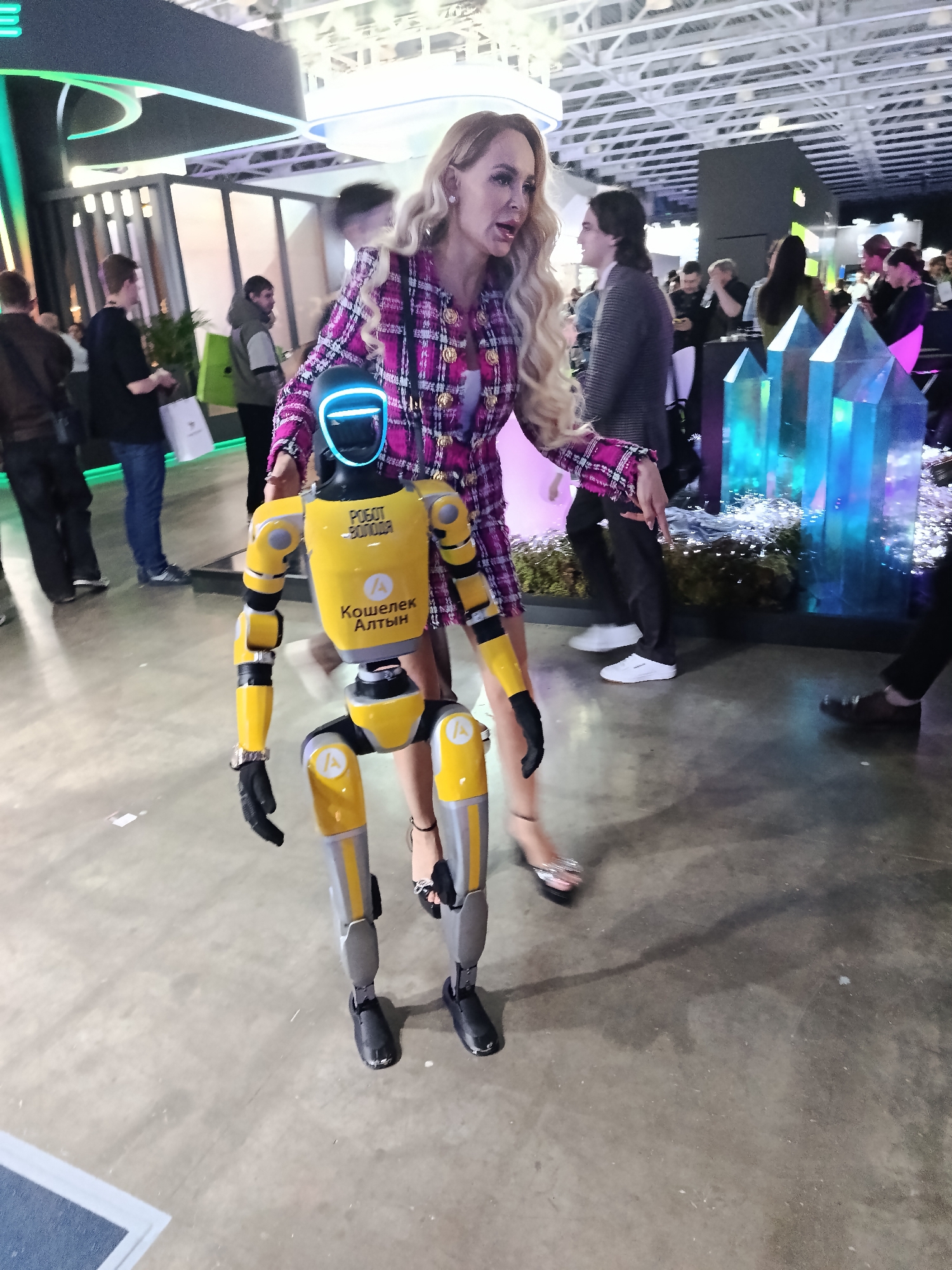 The image captures a scene indoors, possibly at a convention or exhibition in Khimki, Russia. A blonde woman in a pink plaid jacket and skirt is standing next to a yellow and grey robot. The robot has a blue glowing light on its head, and the words "РОБОТ-ВОЛОДЯ" and "Кошелек Алтын" are written on its chest. The woman is wearing silver high heels and a bracelet on her right wrist. In the background, there are numerous people, exhibition displays, and translucent blue crystal-like structures. The floor is a plain concrete surface. The overall environment appears to be a busy, public gathering.
