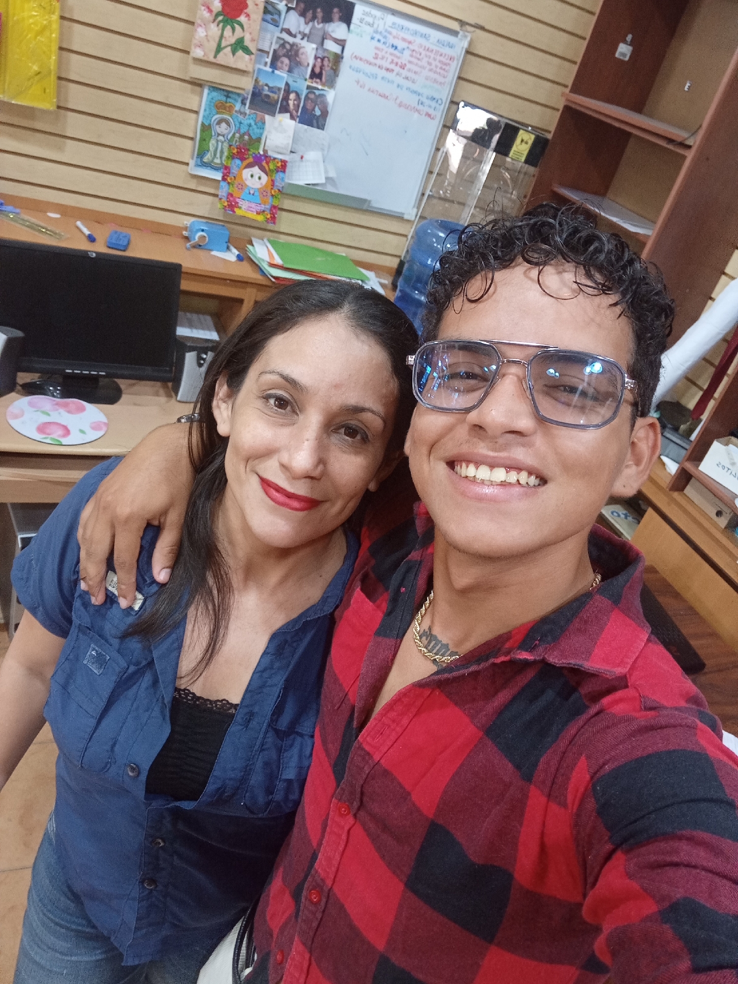 A selfie shows a man and a woman standing close together, possibly inside a commercial establishment in Altagracia, Venezuela. The woman, on the left, has long dark hair, red lipstick, and is wearing a blue button-down shirt over a black top and jeans. Her arm is around the man. The man, on the right, is wearing glasses and a red and black plaid shirt, with a gold chain around his neck. He is smiling. Behind them is a desk with a computer monitor and a white board covered with various papers and images. There are also shelves and a water cooler in the background. The overall environment suggests a casual, indoor setting, likely a small business or office.