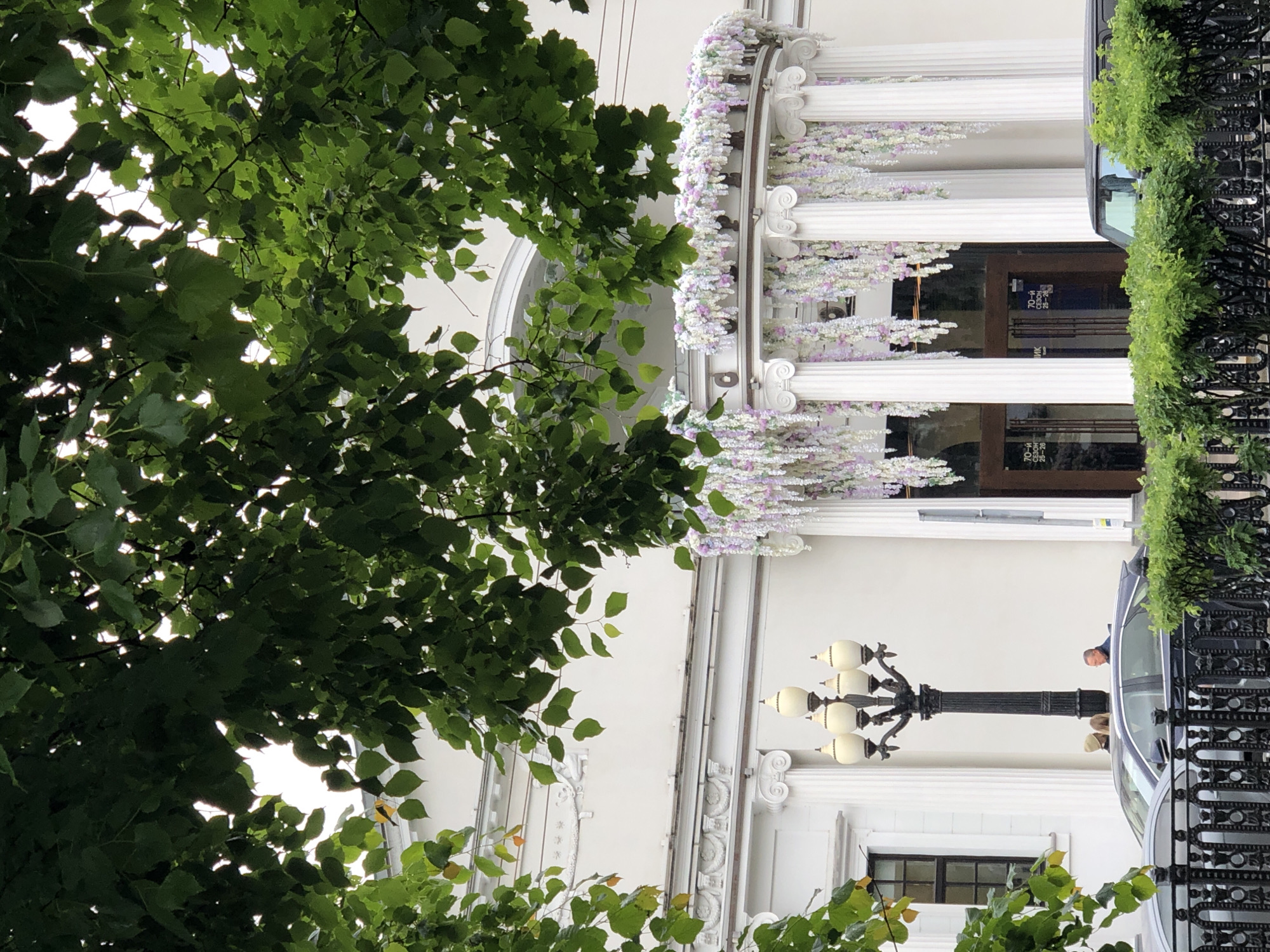 The image shows the facade of a white building with classical columns and ornate decorations, likely in Moscow, Russia. The building is adorned with floral garlands. A black decorative fence runs along the right side, partially obscuring a dark car. A vintage-style lamppost stands in front of the building, and a person is visible in the background near the car. Trees with dense green foliage are prominently featured in the foreground, partially obscuring the view of the building.