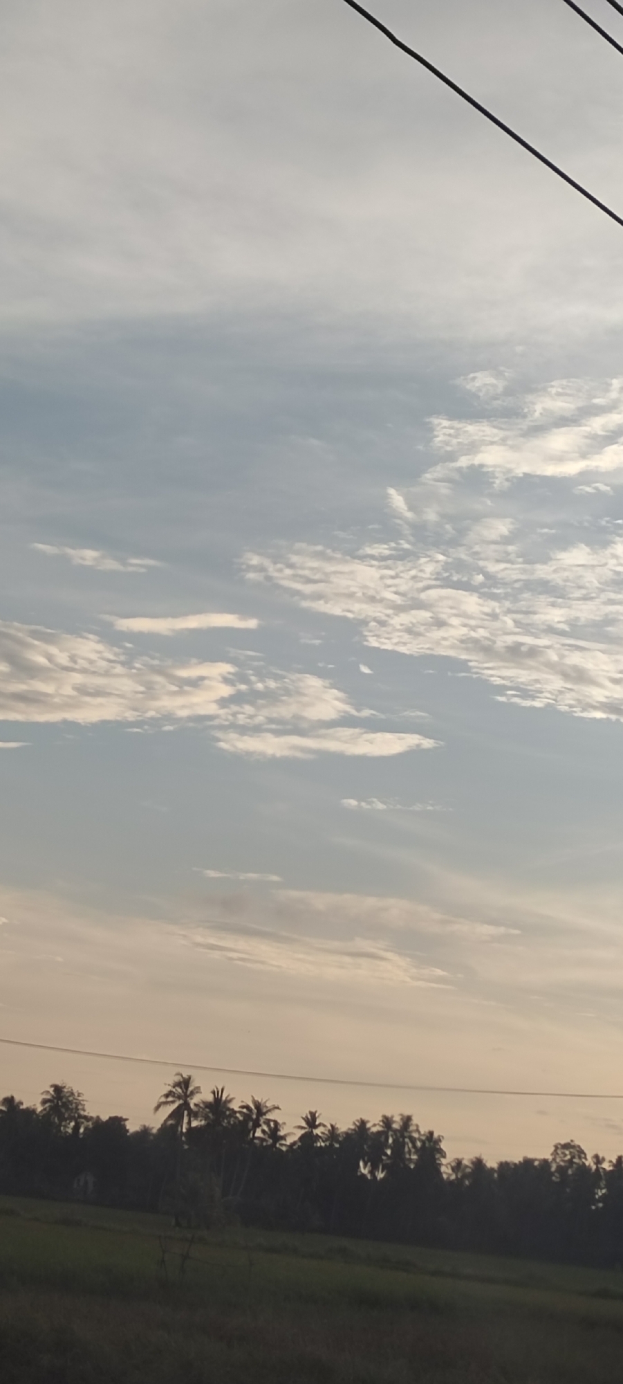 The image depicts a landscape in Kruengseupeng, Indonesia. A field occupies the foreground. In the midground, a line of trees, including palm trees, stretches across the horizon. Overhead, the sky is filled with light-colored clouds. Two dark, diagonal power lines extend from the top right corner.