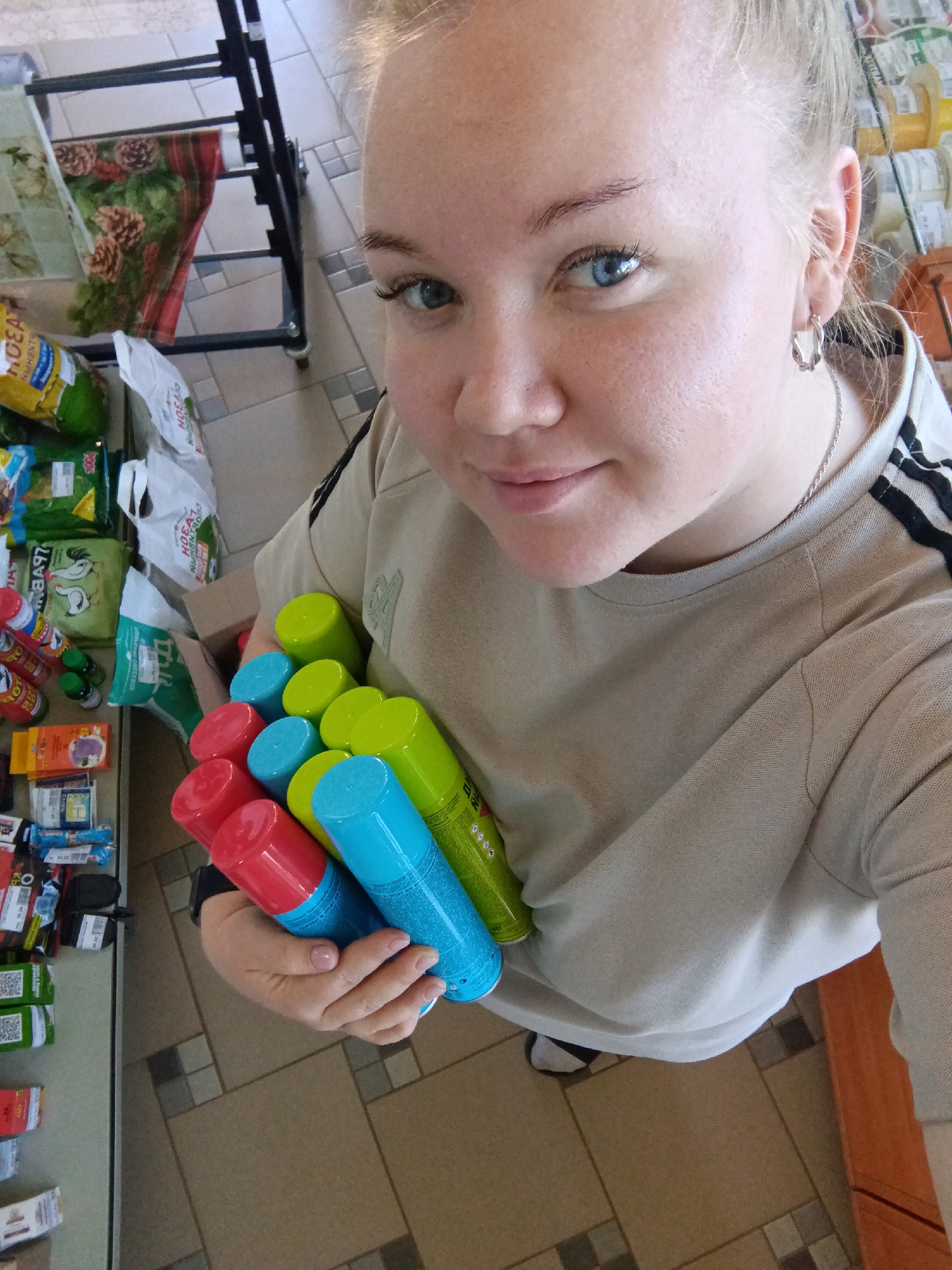 A woman is holding several cans of spray paint in her left arm. The cans are red, blue, and green. She is wearing a light brown t-shirt with black stripes on the sleeves and has light skin, blue eyes, and blond hair pulled back. She is wearing a silver chain necklace and small silver hoop earrings. She appears to be in a store, as shelves of products are visible to the left and behind her. The floor is tiled in a geometric pattern. The location could be Фоки, Russia, given the context of the prompt.