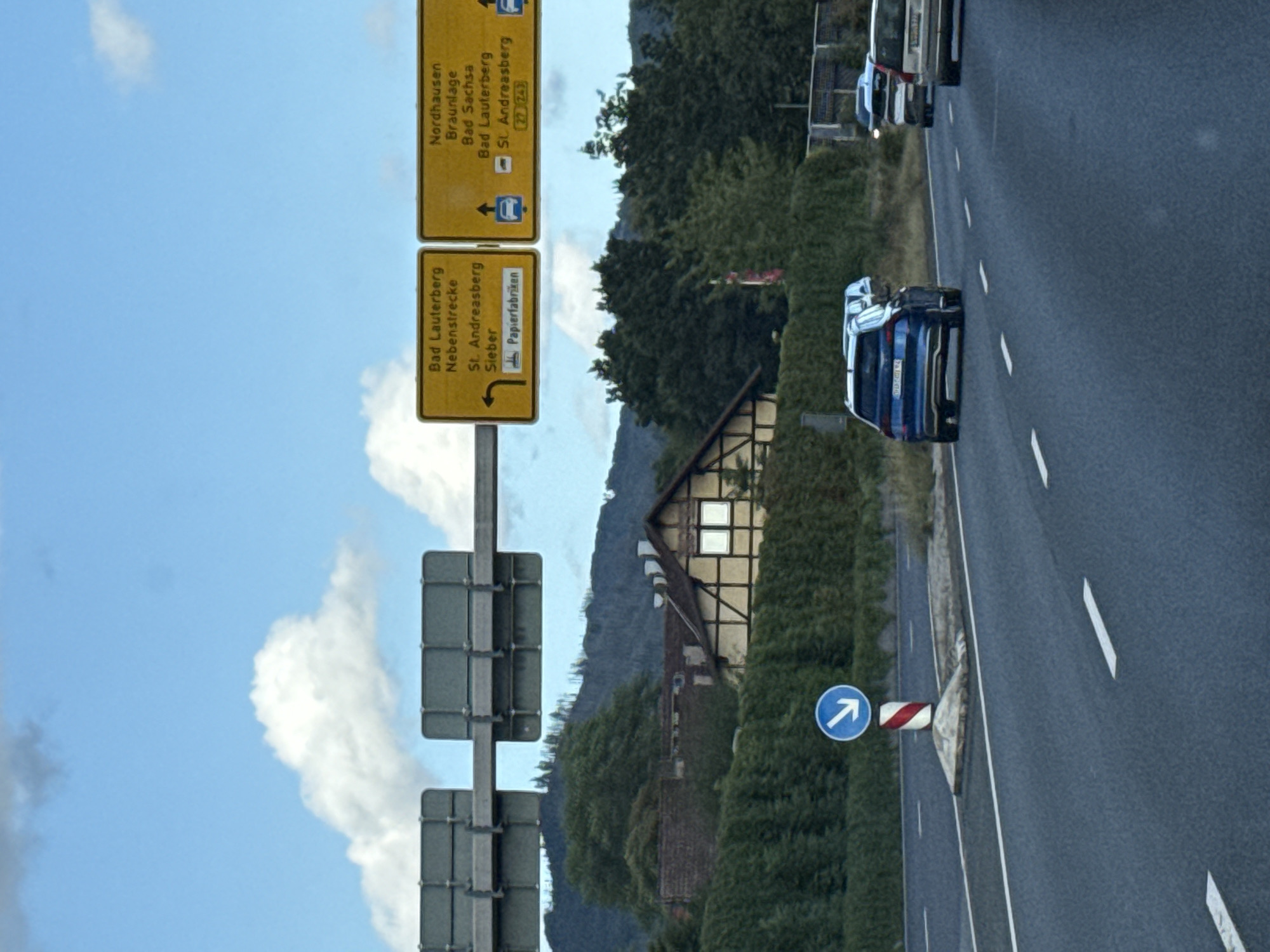 The image shows a highway scene with several cars driving on a paved road. In the background, there is a lush green hedge, a traditional German half-timbered house, and a forested hillside under a partly cloudy sky. Overhead, traffic signs direct drivers to destinations such as "Bad Lauterberg," "Nordhausen," and "Braunlage." A blue sign with a white arrow indicates a turn. The overall scene suggests a rural or suburban setting in Germany, likely near Herzberg.