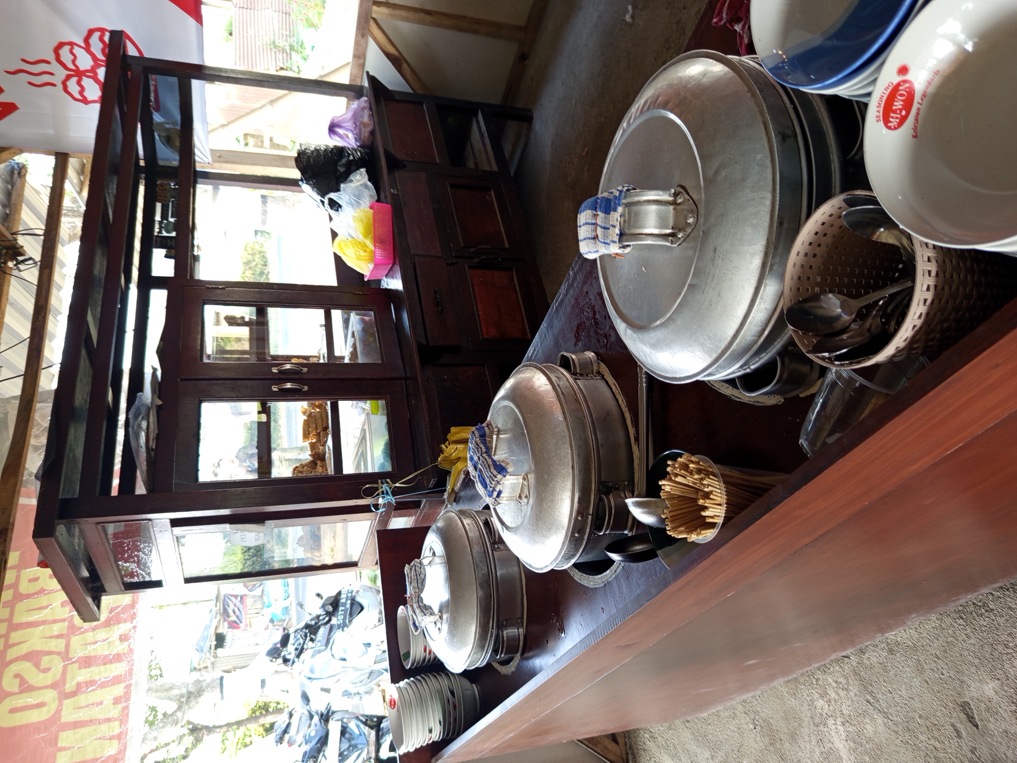 This image depicts an outdoor food stall in Bokor, Indonesia. The stall features a wooden counter with several large, aluminum steaming pots. A stack of white plates is visible on the left side of the counter. Various utensils, including spoons, are contained in a woven basket. Several plastic bowls, some stacked, are also present. In the background, a wooden cabinet with glass doors displays items, and a red sign with yellow lettering is visible. Motorcycles are parked to the left of the stall, suggesting it is located near a road. The ground is a mix of concrete and dirt.
