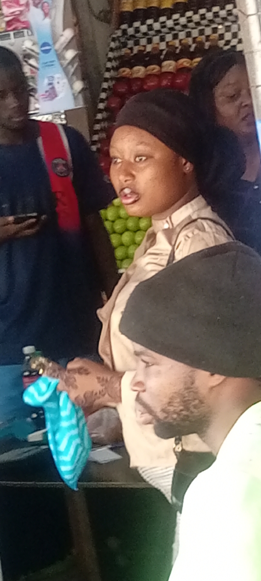 The image appears to be taken outdoors, possibly at a market stall in Aviation Quarters, Nigeria. There are several people present. A man with a black hat and beard is in the foreground, partially obscuring the view. A woman wearing a black head covering and a light-colored shirt is looking to the left with an expression of surprise or excitement. Another man is wearing a navy shirt with a red and black strap. In the background, a woman with a dark head covering is visible. A display of round, green objects (possibly limes or small green fruits) can be seen behind the woman in the light-colored shirt. To the left is a bottle of what appears to be coke. The background also shows a display of items, including possible beverages and packaged goods. The stall's surface appears to be black, and a folded blue cloth is on it.