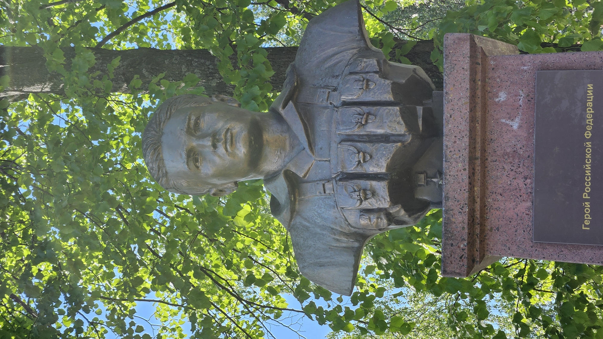 A bronze bust of a young, short-haired male, rendered with a serious expression, is centered in the image. The figure wears a collared uniform top and a tactical chest rig equipped with at least five visible pouches across the front. The bust is mounted on a multi-tiered pedestal made of reddish-brown speckled stone. A dark rectangular plaque, inscribed with gold Cyrillic text translating to "Hero of the Russian Federation," is affixed to the lower front of the pedestal. The background is dominated by a dense canopy of vibrant green deciduous tree leaves, illuminated by bright sunlight, against a clear blue sky. A tree trunk is visible behind the bust's right side. The scene is outdoors, well-lit, and appears to be in a park or memorial area within the city of Unknown, Unknown.