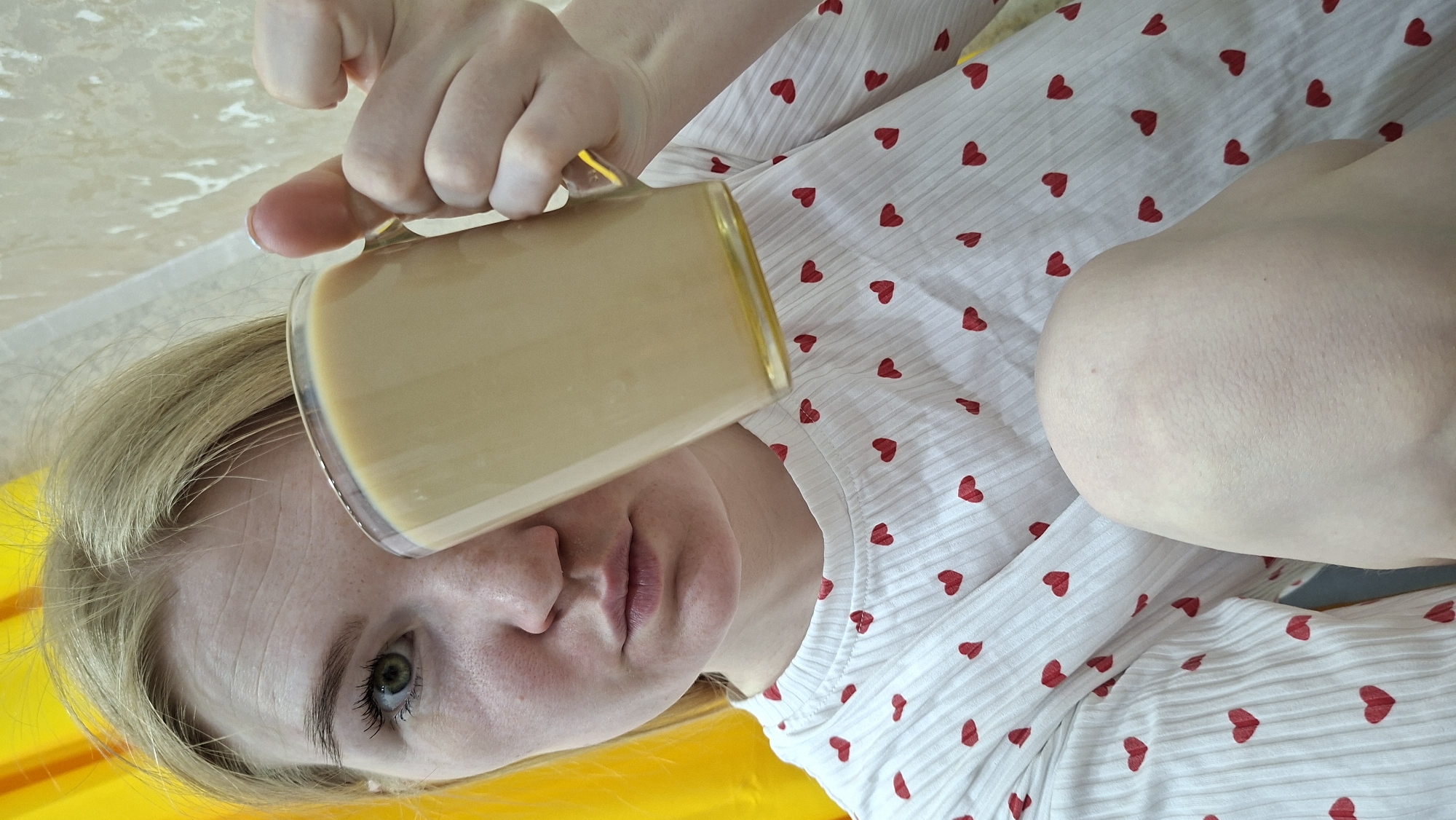 A fair-skinned woman lies on a white fabric surface patterned with small red hearts, holding a clear glass mug filled with a light brown liquid. Her blond hair is visible under the mug. She is indoors. A yellow surface is partially visible. The location is Chaikovsky, Russia.