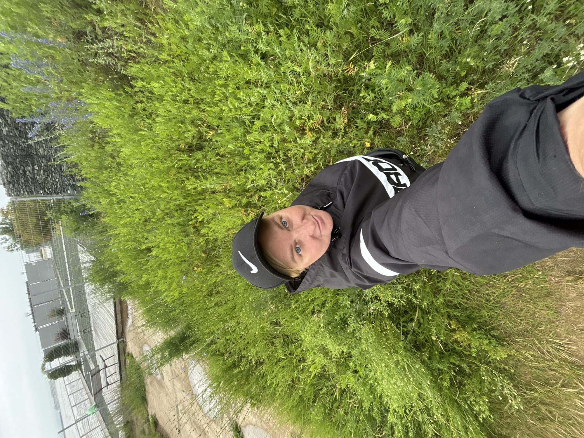 A person is lying down in a grassy area, facing the camera. They are wearing a black baseball cap with a white Nike logo on it, and a black jacket with white stripes on the sleeves. The person has fair skin and blue eyes, and they are smiling. The background is a lush green field with some small purple flowers. To the left, there is a glimpse of a grey, fence-like structure and a body of water, suggesting the person is near an outdoor facility or park by a body of water. The image is taken from a low angle, looking up at the person. The lighting suggests it is daytime, possibly overcast due to the muted tones. The overall impression is casual and relaxed.