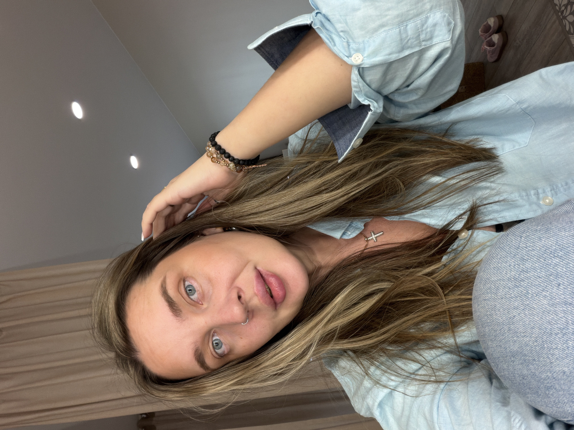 A woman with light brown hair and blue eyes is pictured indoors. She is wearing a light blue button-down shirt and has several bracelets on her left wrist. A cross pendant is visible around her neck. Her hair is styled with highlights and layers, and she has a ring piercing in her left nostril. The background features a plain, light-colored wall with ceiling lights. A pair of slippers are partially visible in the upper right corner. The location is possibly Moscow, Russia, but no specific landmarks or identifiable indicators are present to confirm the location within the city.