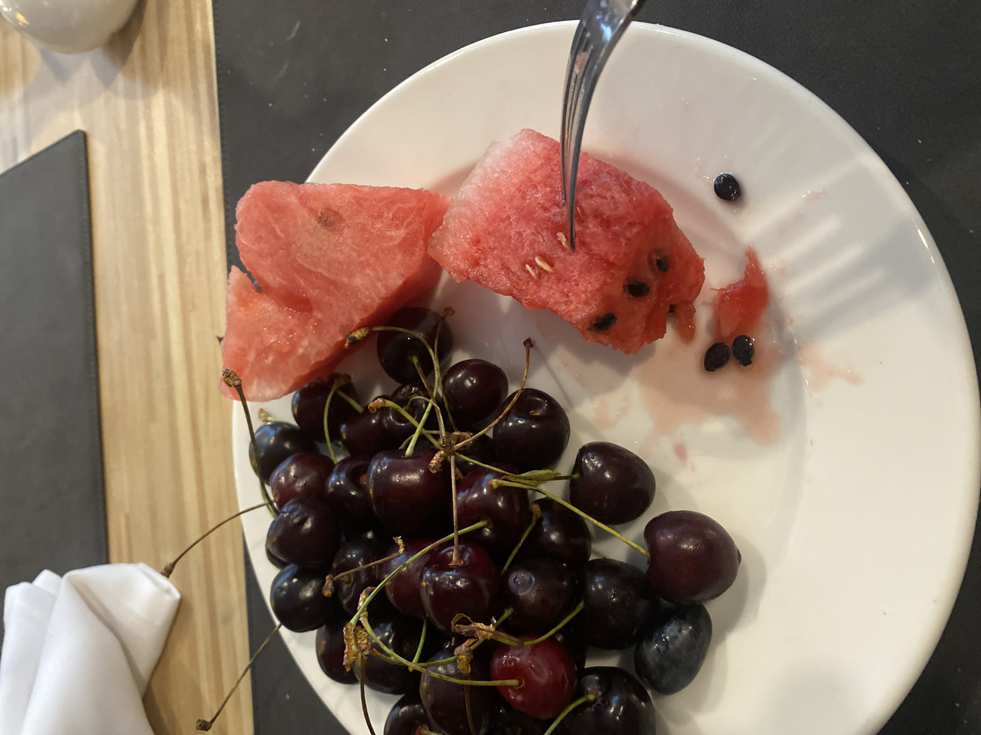 A close-up, eye-level shot shows a white plate with a serving of cherries and watermelon. The cherries, dark red and plump, are piled on the left side of the plate, some with stems still attached. Two pieces of watermelon, cut into wedge shapes and one with a bite taken out of it, are positioned on the right. A silver fork is shown piercing one of the watermelon pieces, suggesting someone is about to eat it. A white napkin is folded and placed behind the cherries on the upper left of the plate. The plate sits on a dark surface, possibly a placemat, which is on top of a wooden table. The background is blurred, showing a light wood grain and a portion of a white rounded object. The overall scene is clean and appears to be a meal setting, though no specific location within the city of Aksu, Turkey, can be determined from the visual information provided.