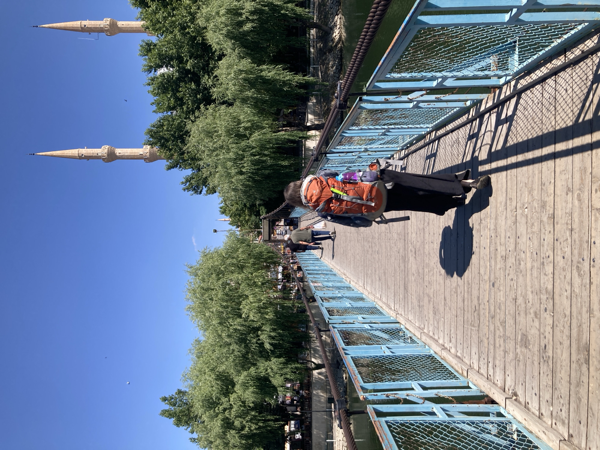 A sunny day scene captures a pedestrian bridge over a body of water, likely the Kızılırmak River in Avanos İlçe Merkezi, Turkey. The bridge features a weathered wooden deck and light blue metal railings with diamond-shaped wire mesh. Several people are crossing the bridge. In the foreground, a person with an orange backpack walks away from the camera, their shadow cast on the wooden planks. Behind them, other individuals are also walking across the bridge. Lush green trees line the banks of the water. In the background, two minarets of a mosque rise above the trees, indicating the presence of religious architecture. The sky is a clear, vibrant blue, with a few distant birds or small aircraft visible. The overall impression is one of a pleasant outdoor activity in a scenic, culturally rich location.